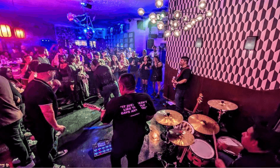 People dancing and socializing in a lively nightclub with colorful lighting and a drum set in the foreground.