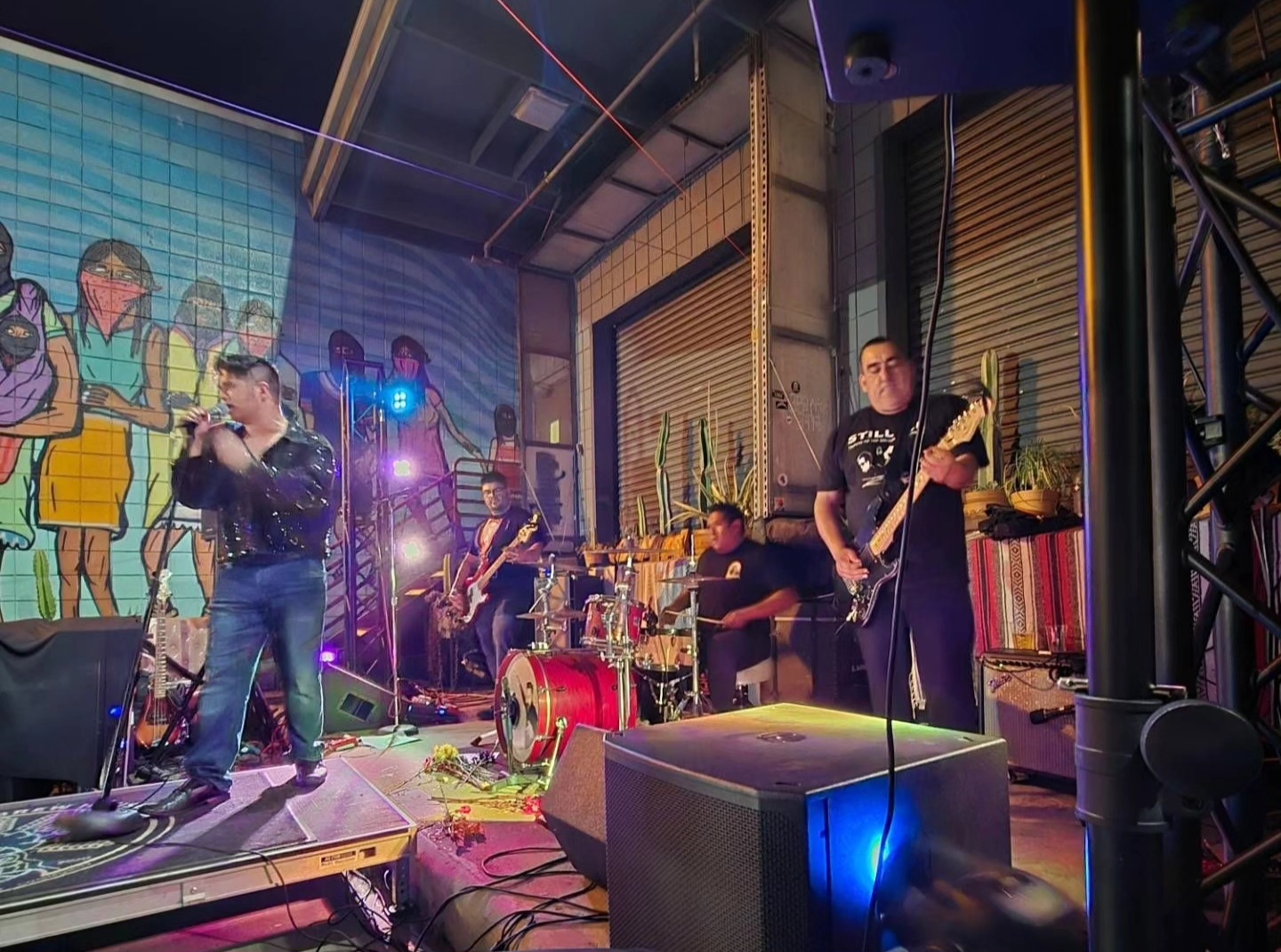 Band performing on stage with colorful background, musical instruments, and stage lighting, in an indoor venue.