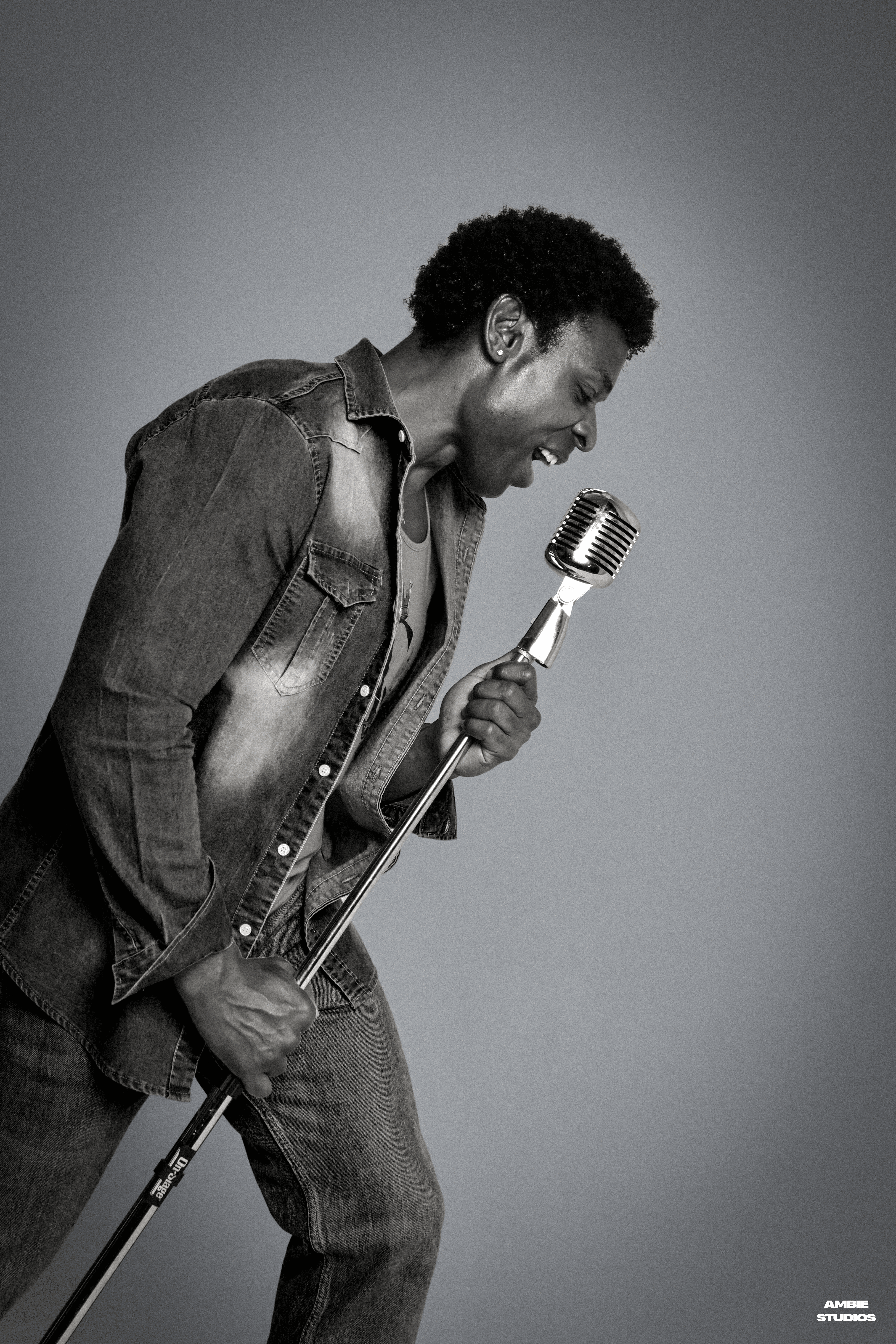 Man singing into a vintage microphone, holding the stand, wearing a denim jacket, in black and white.