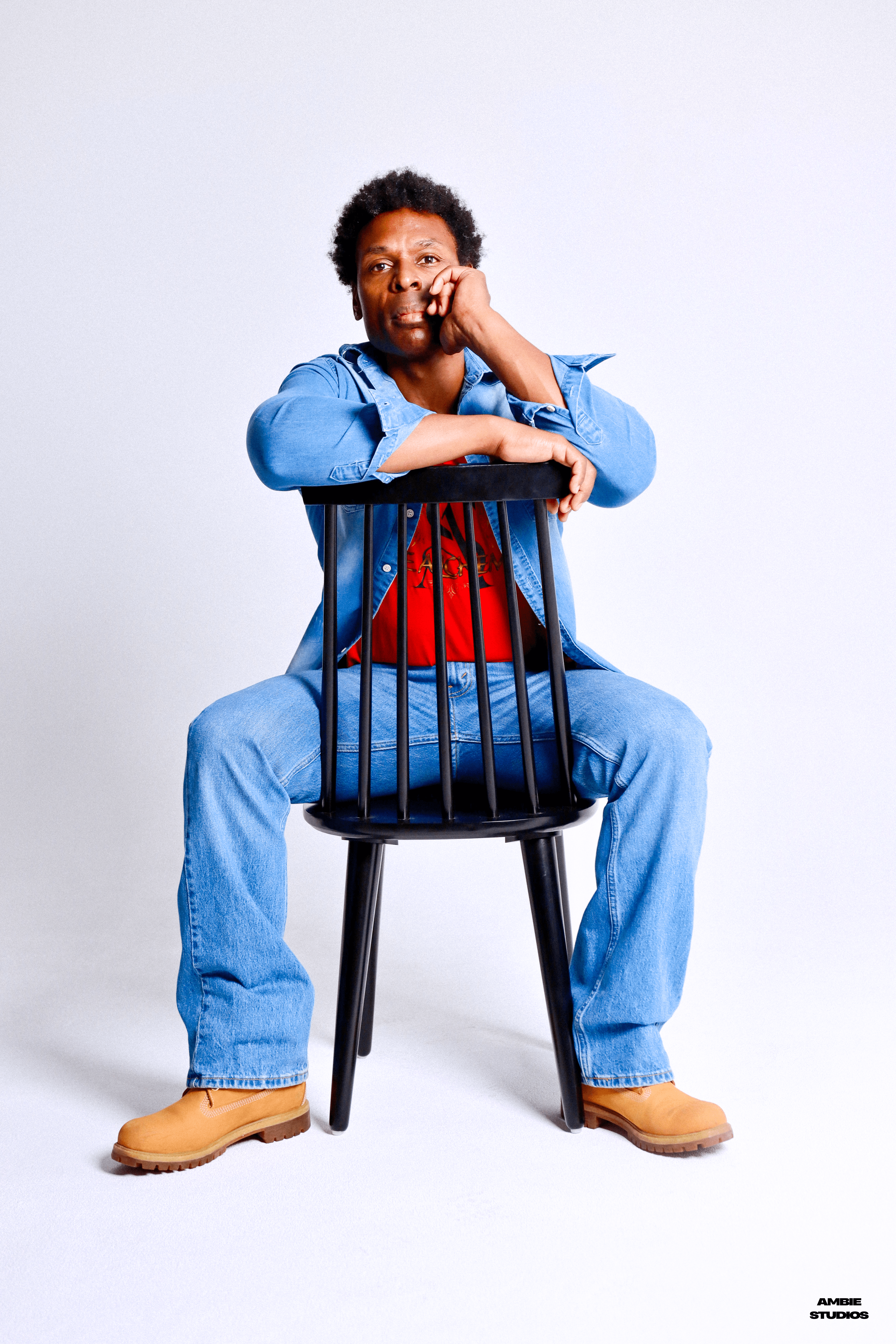 Person sitting on a chair, smiling, with one hand on their face, wearing a blue jacket, red shirt, and jeans.