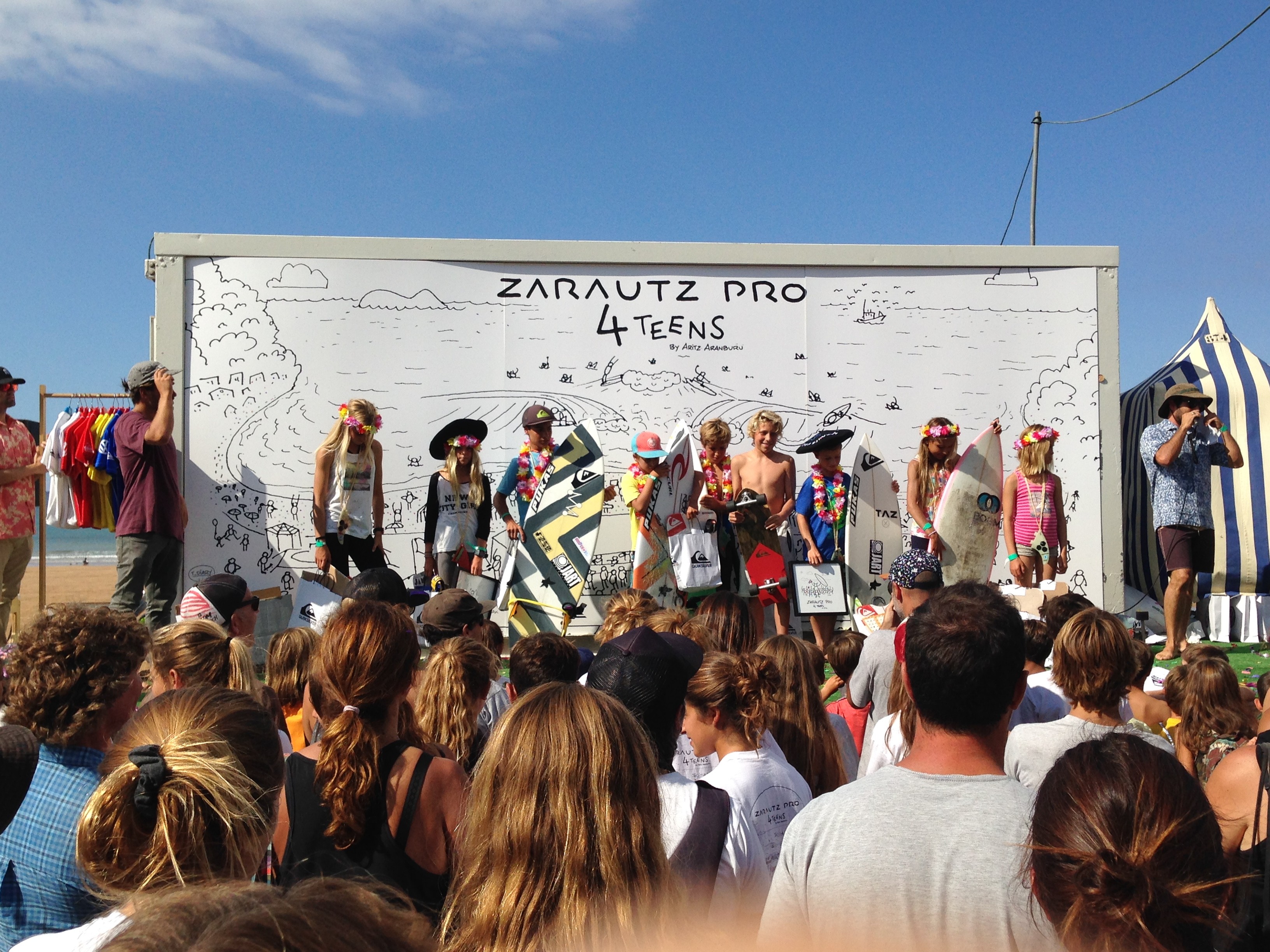 Group of children and adults on stage during a performance at an outdoor event, with a crowd watching in front.