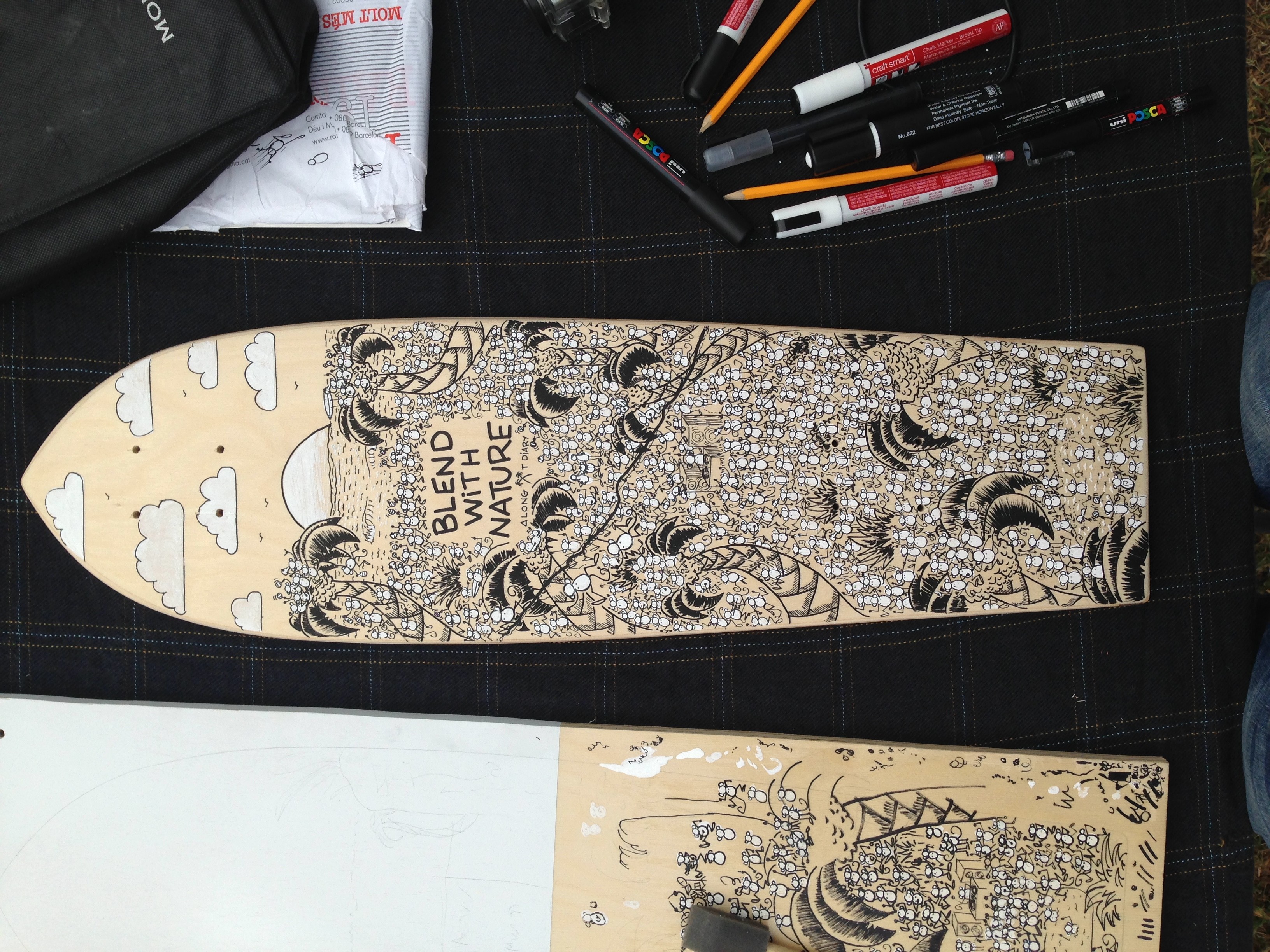 A skateboard deck with a detailed black and white illustration, surrounded by markers and art supplies, on a dark surface.