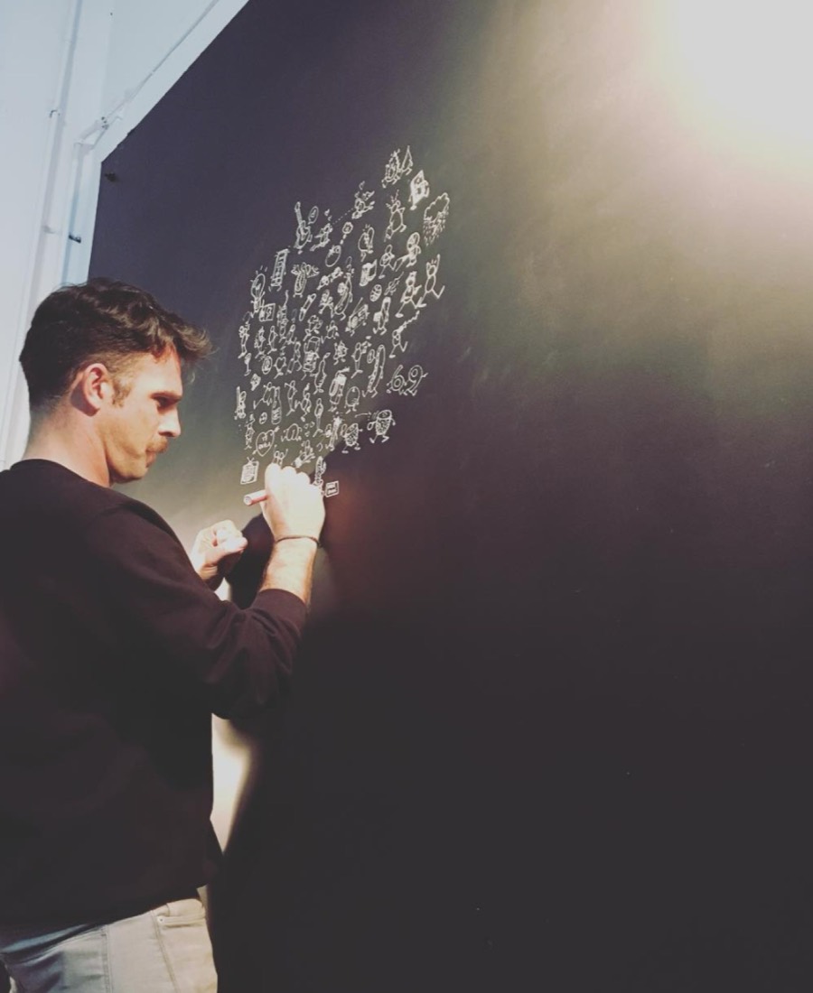 Person writing on a black wall with white chalk, creating a tree shape with words or symbols, in a well-lit room.