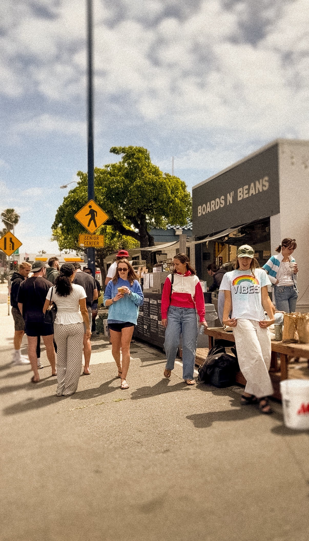 People gather outside a building with a sign reading 'Boards n Beans' on a sunny day.