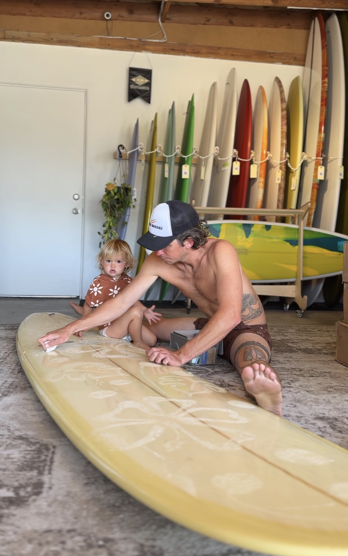 Young man and child on a surfboard indoors, with surfboards and paddles in the background.