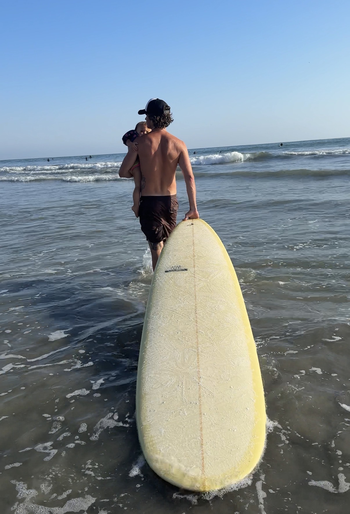 Person carrying a surfboard walking into the ocean at the beach, with another person nearby, under a clear blue sky.