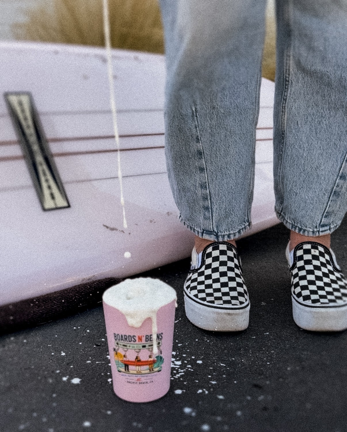 Person wearing checkered slip-on shoes and jeans standing next to a pink cup with a cartoon design, on a dark surface.