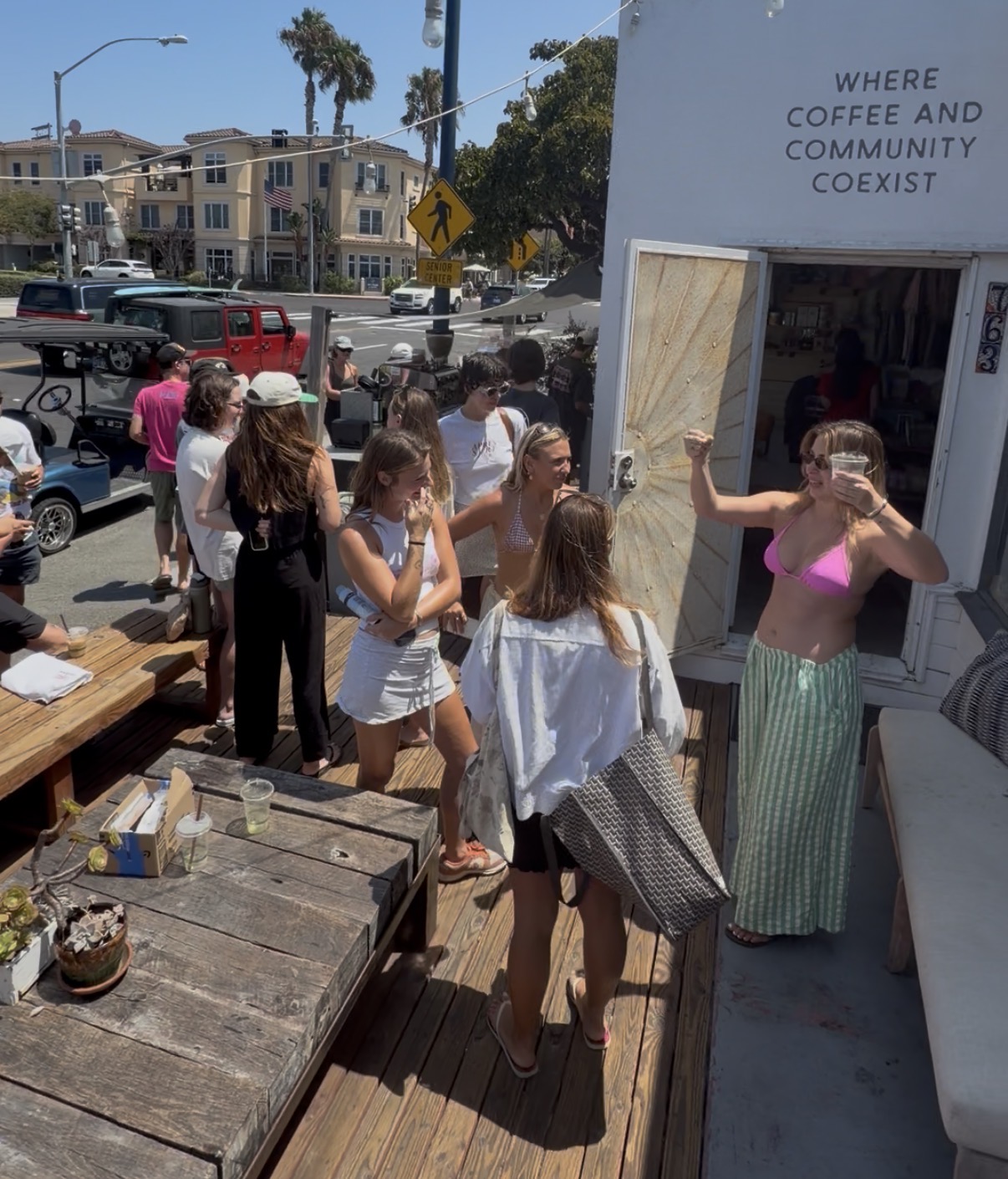 Group of people waiting outside a coffee shop, some talking and others looking at the shop, sunny day, buildings in background.