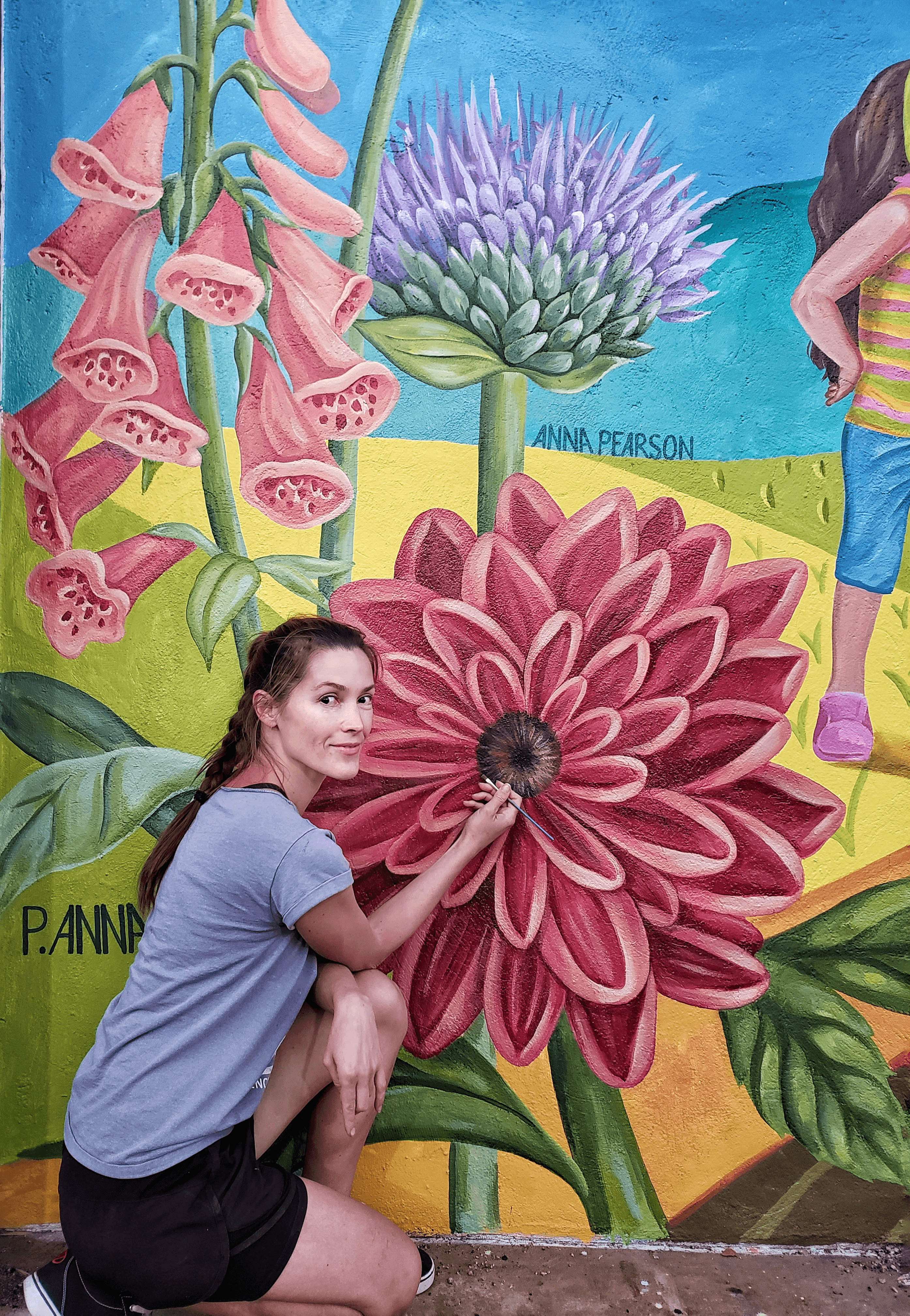 Young woman crouching in front of a colorful mural with large flowers and a person in striped clothing in the background.