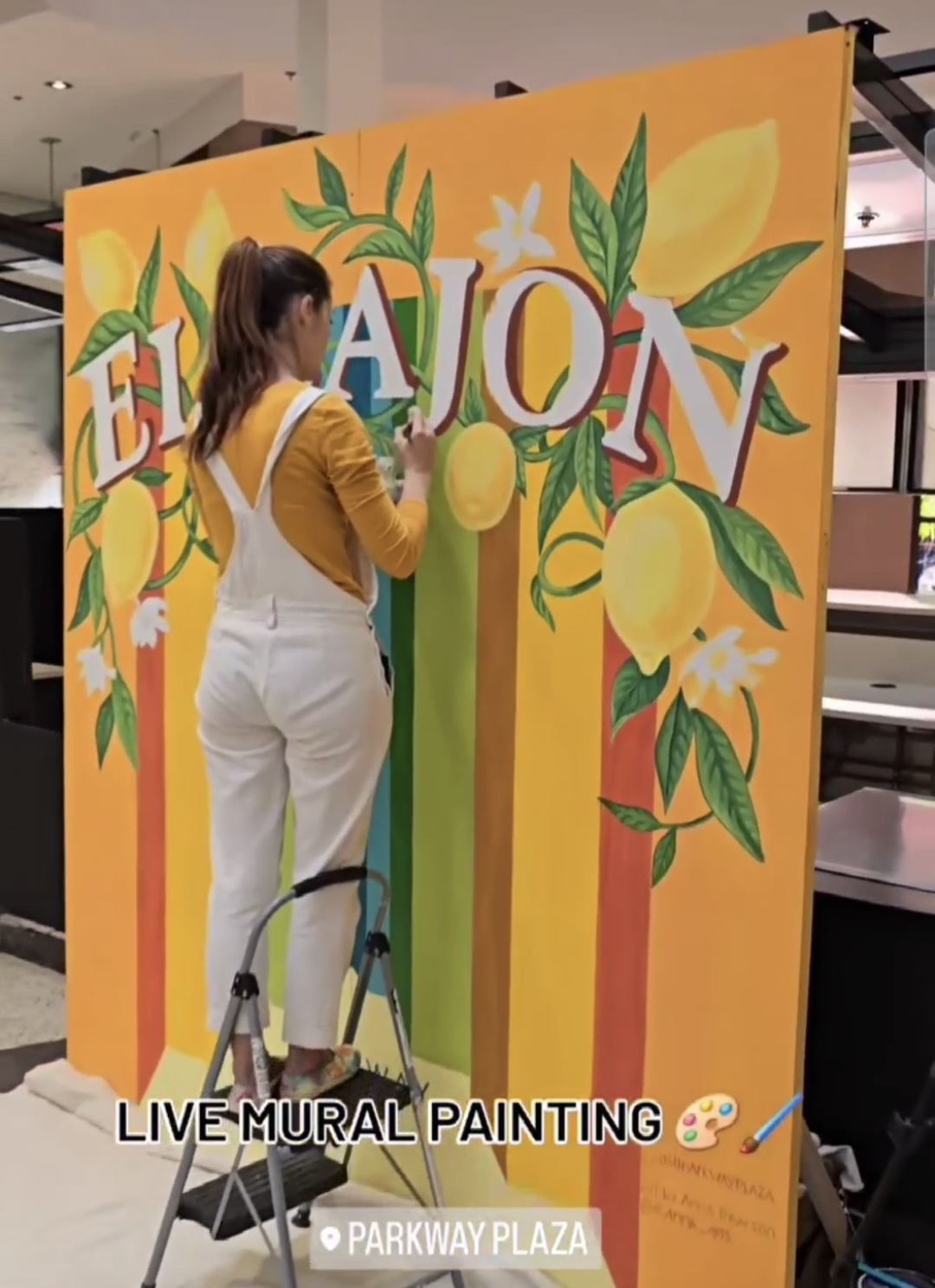 Person painting a colorful mural with lemons and leaves, standing on a ladder, with text 'LIVE MURAL PAINTING' and location details.