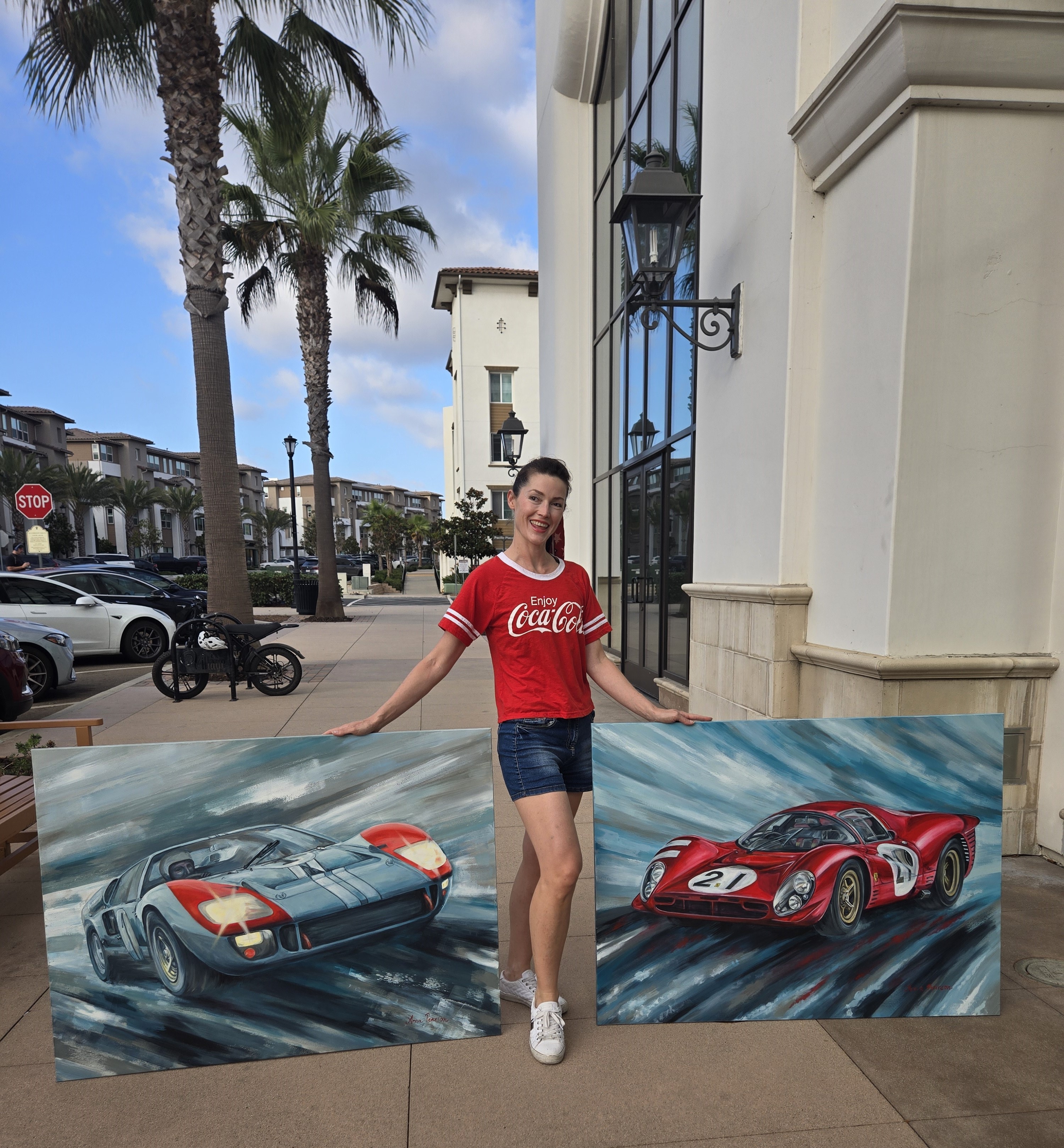 Young woman standing between two racing car paintings outside building, palm trees in background, sunny day.