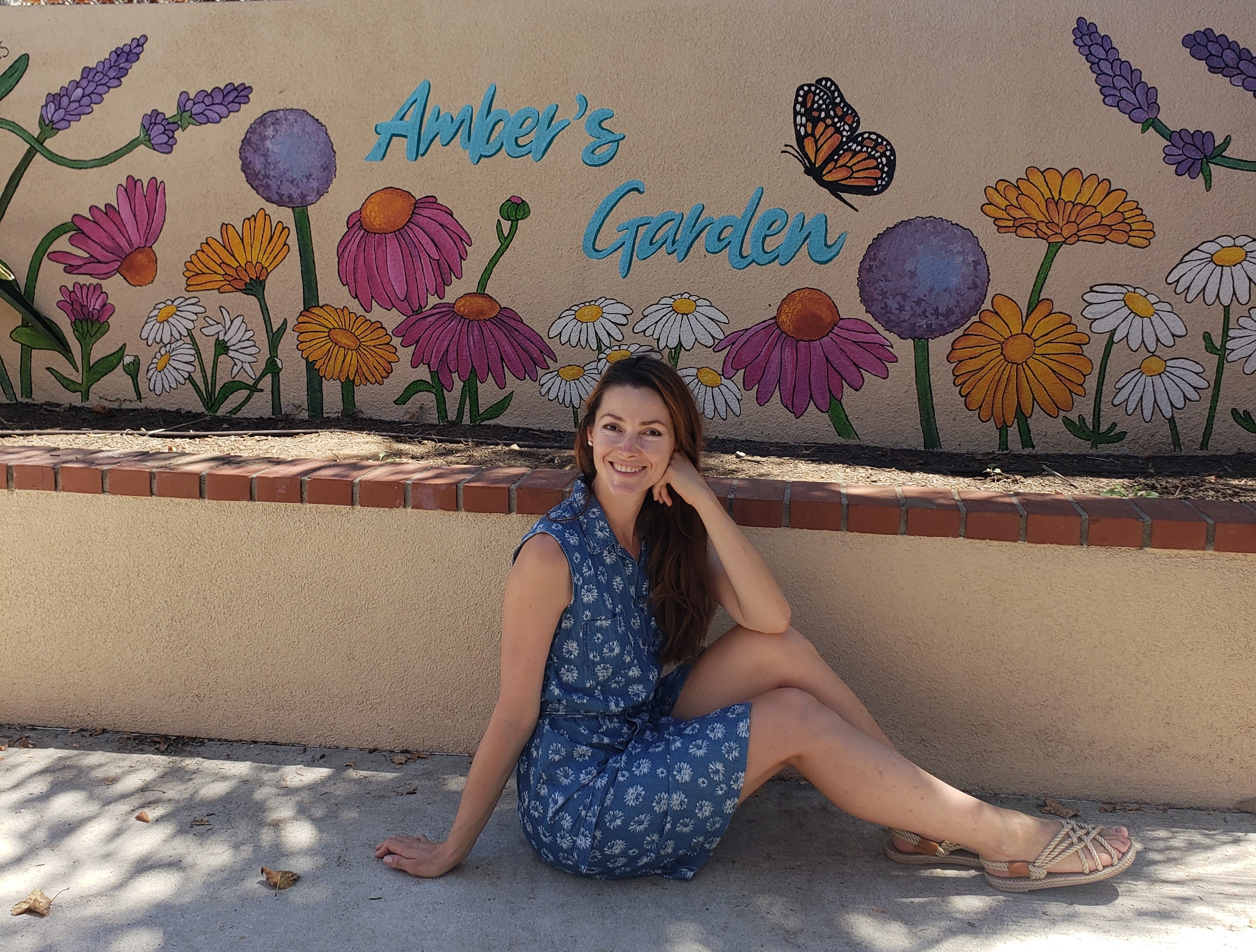 Young woman sitting on ground in front of a colorful mural with flowers and a butterfly, smiling, wearing a blue dress.