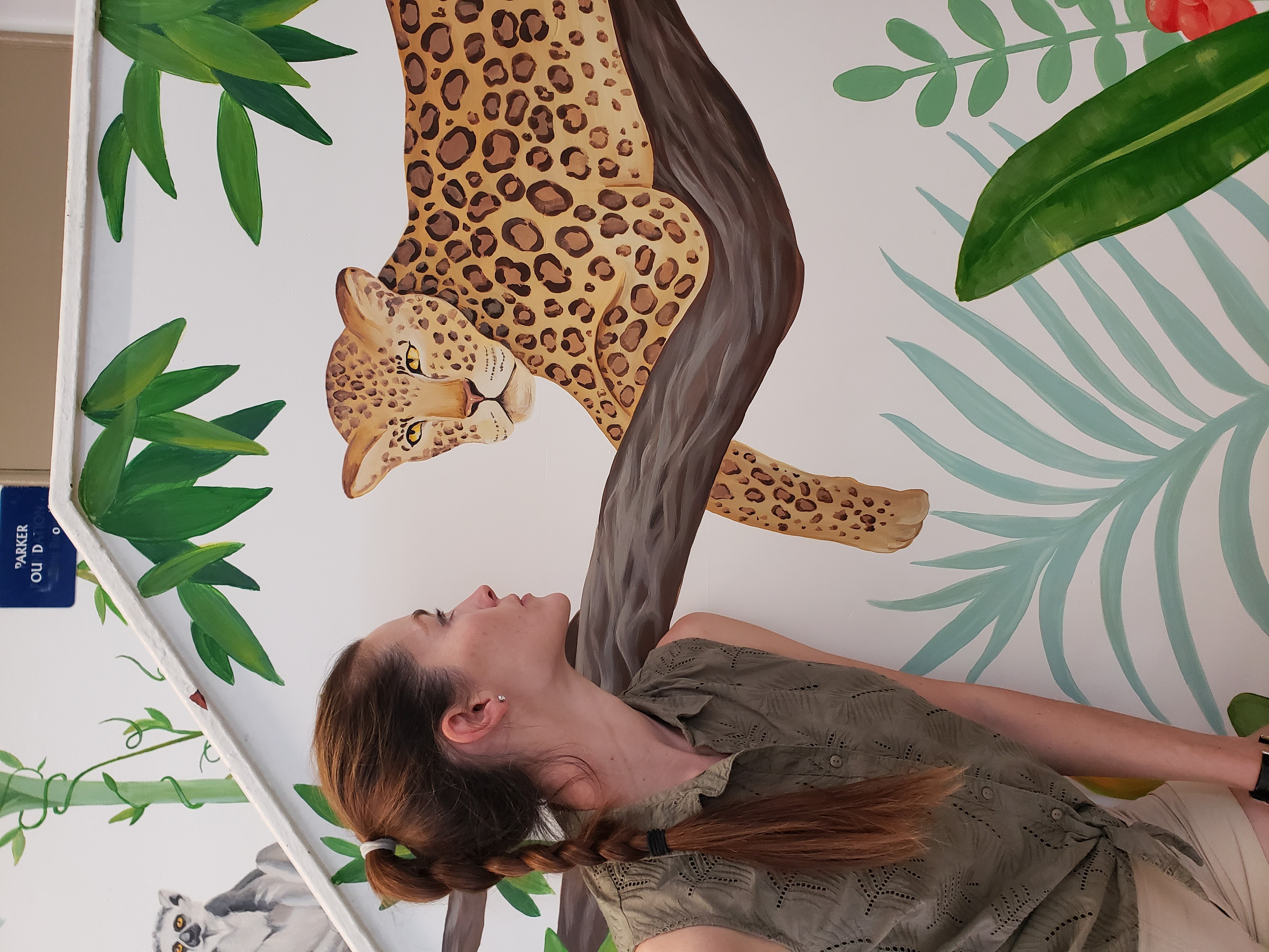 Person with long hair looking up at a mural of a leopard in a jungle setting with green leaves.