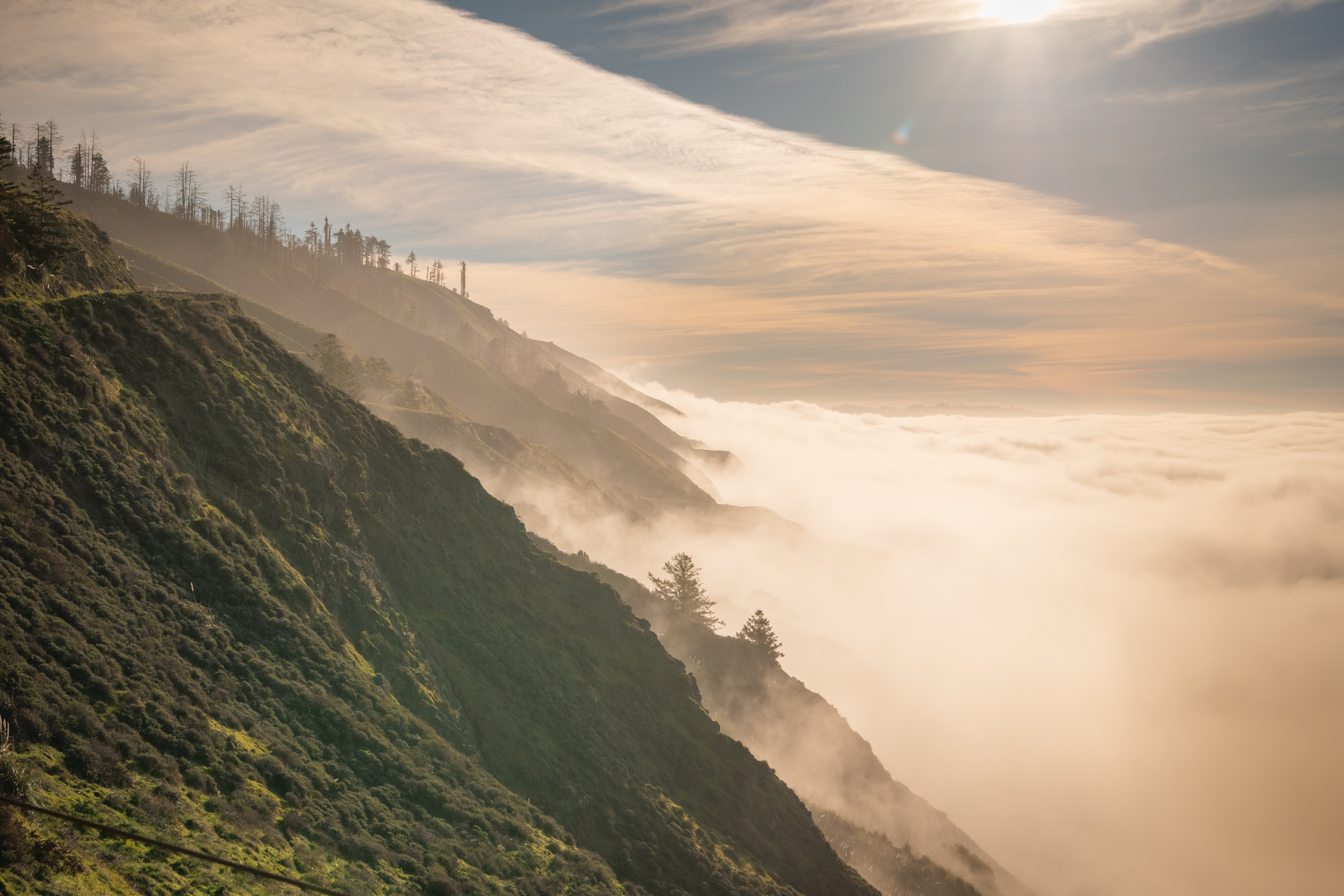Mountain slope covered with greenery, fog or clouds below, sky with clouds and sunlight, distant trees on ridge.