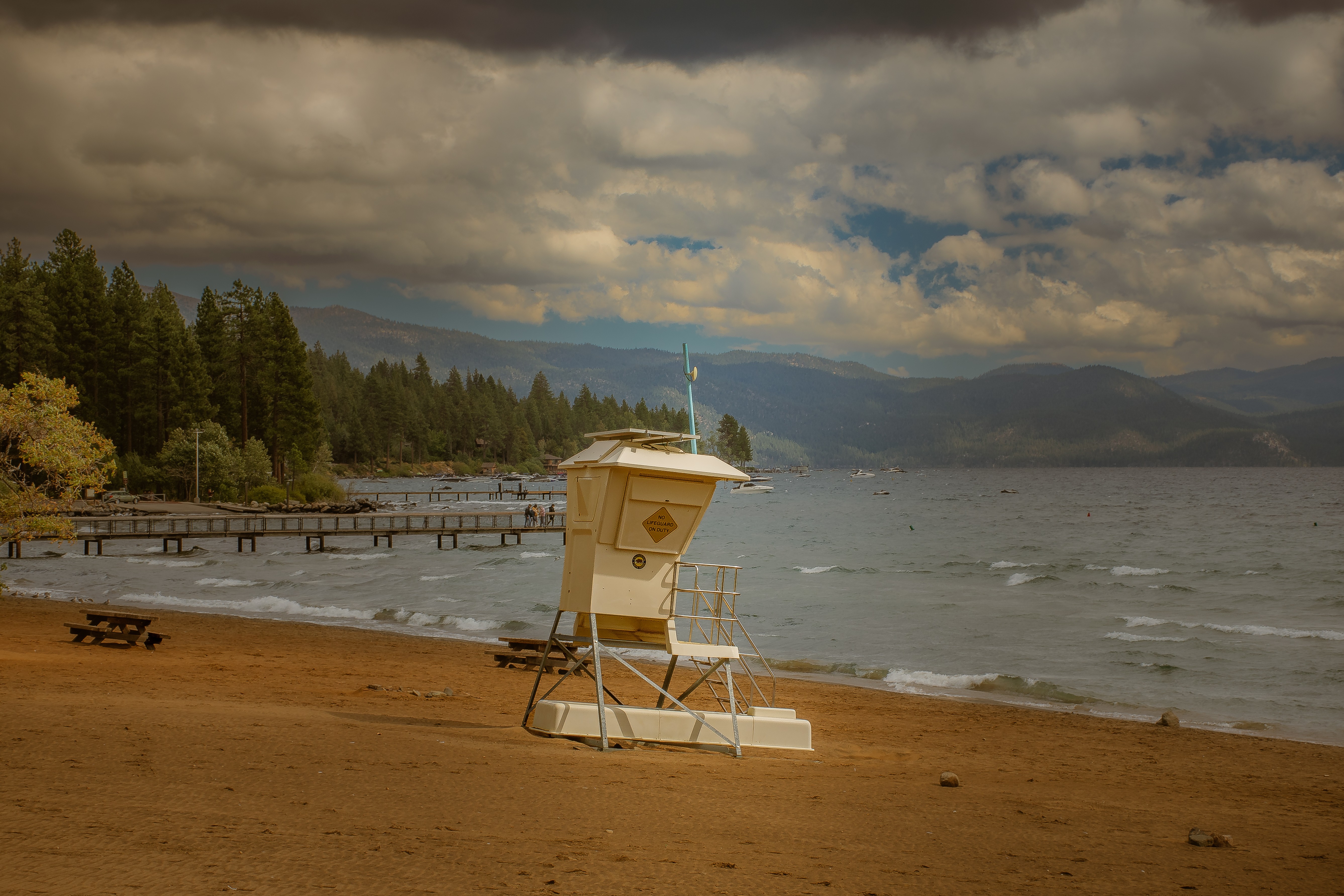 Lifeguard tower on a sandy beach with trees and mountains in the background, cloudy sky, and calm water.