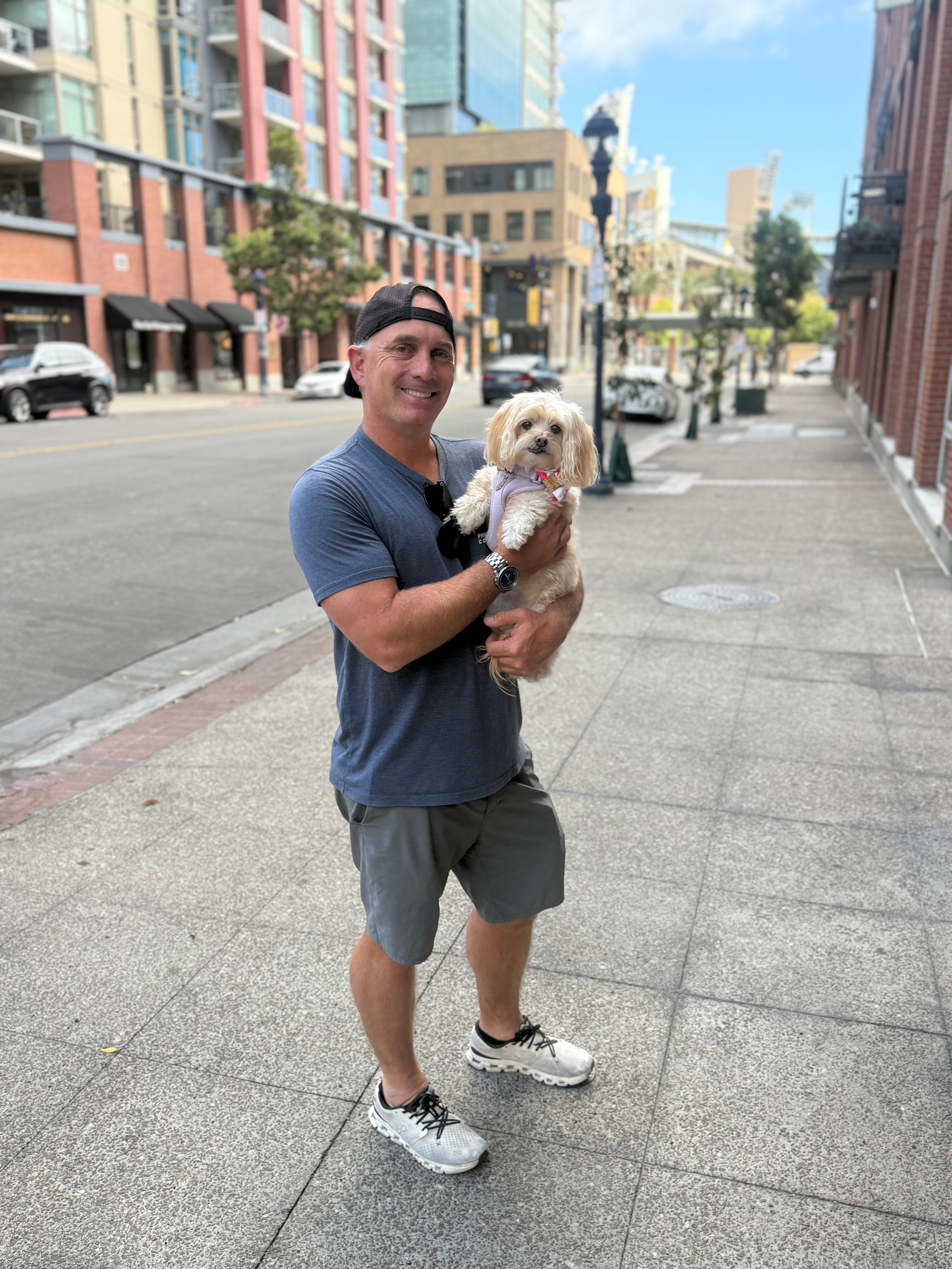 Young man holding a small dog on a city sidewalk, buildings and trees in the background, sunny day.