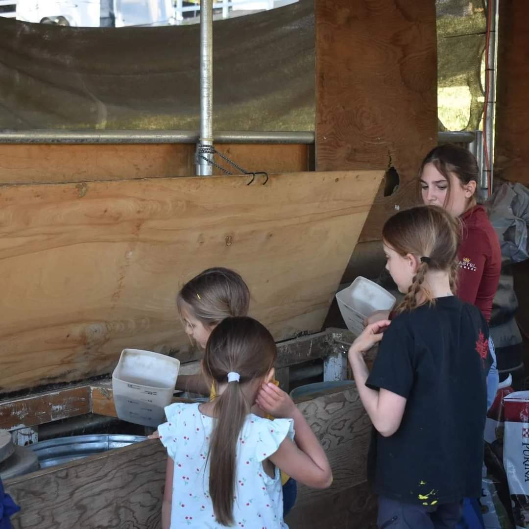 Four children with braided hair and a woman look at a display or exhibit indoors, with wooden and metal structures in the background.