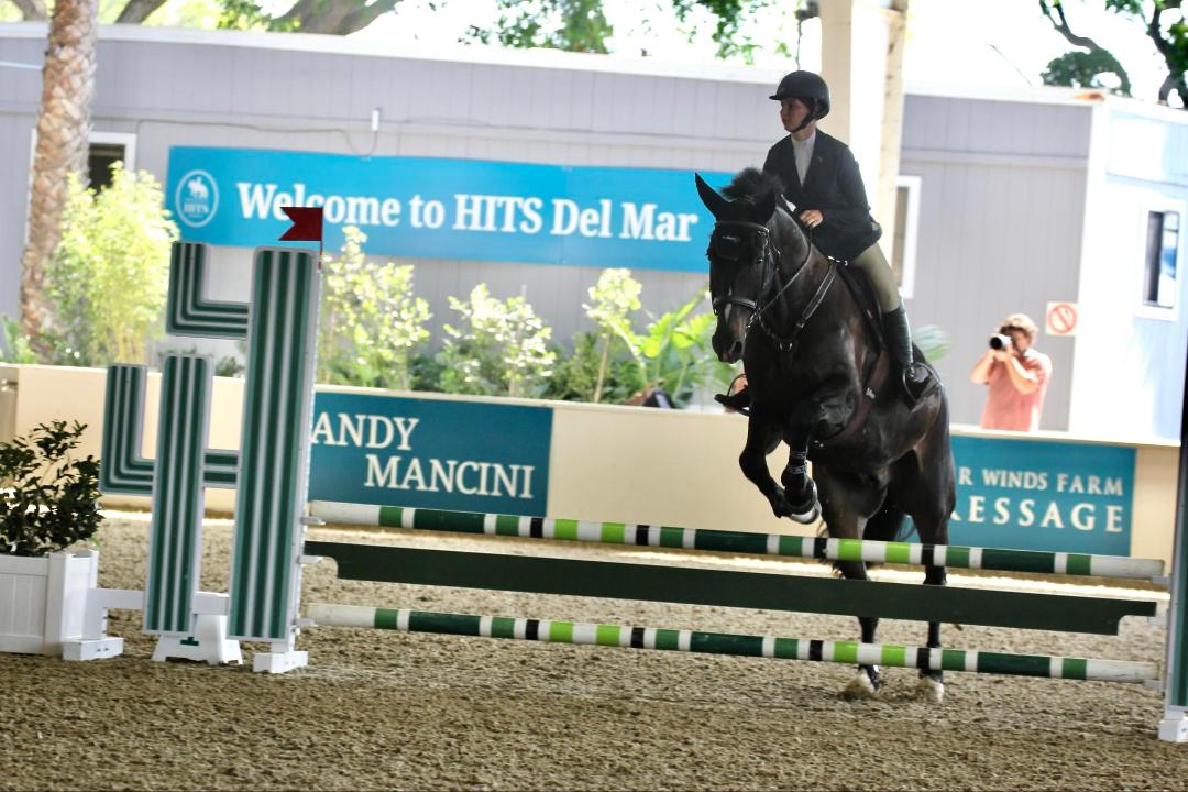 Person riding a horse jumping over an obstacle in an indoor arena, with a sign in the background reading 'Welcome to HIT'S Del Mar'.