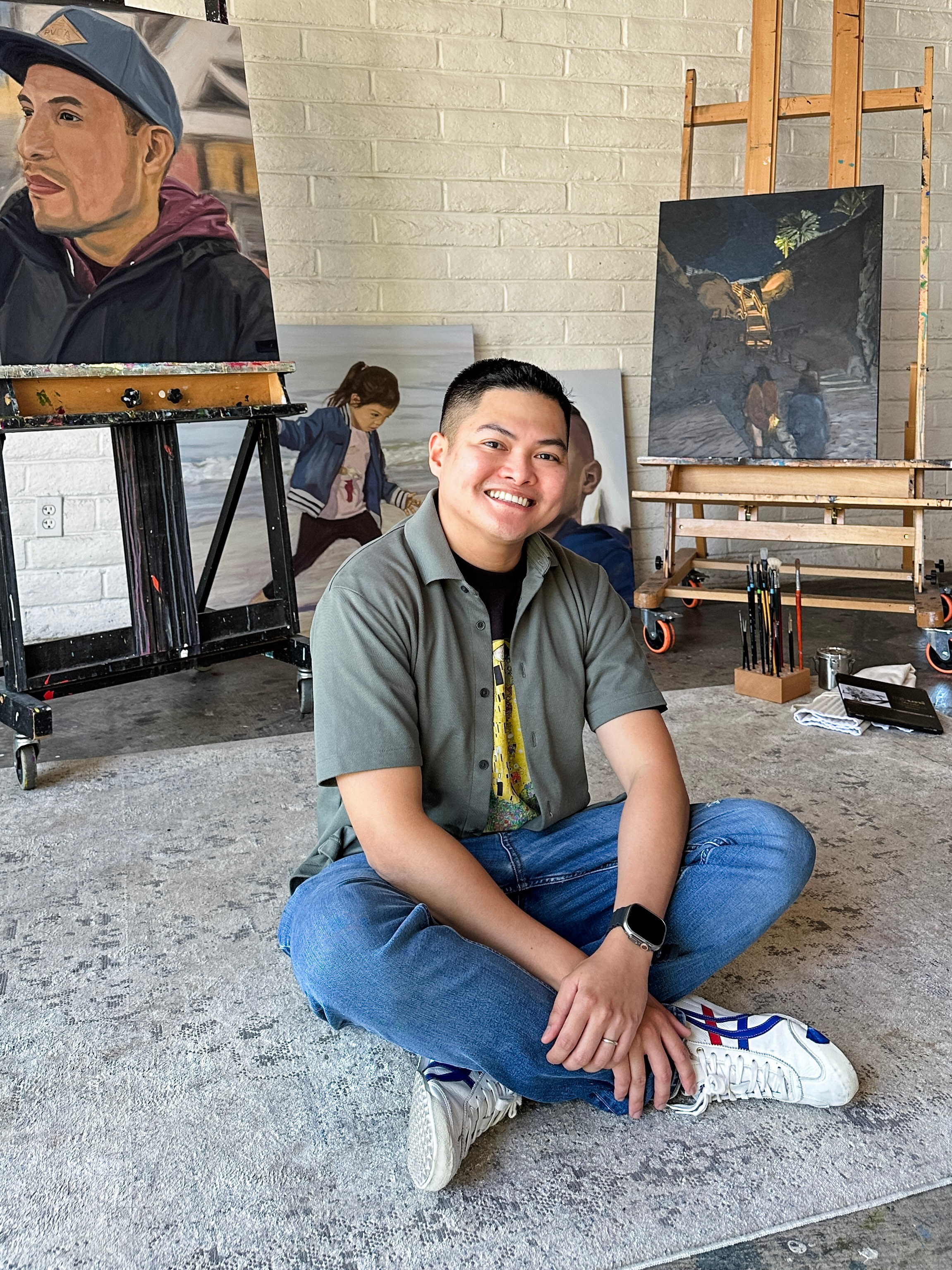 Young man sitting cross-legged on the floor, smiling, in an art studio with paintings and easels behind him.