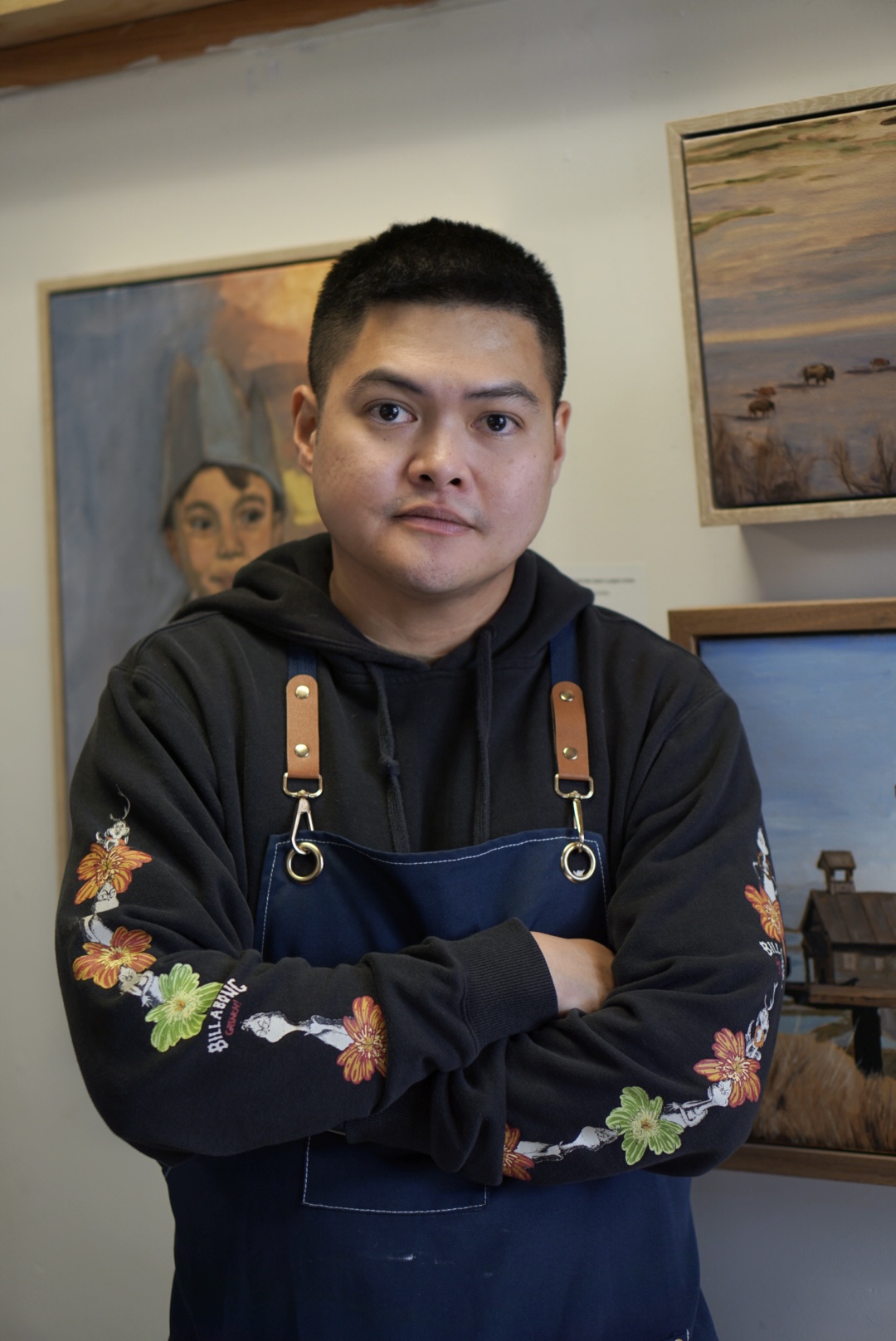 Young man with short black hair wearing a black hoodie and blue apron, standing with arms crossed in front of paintings.