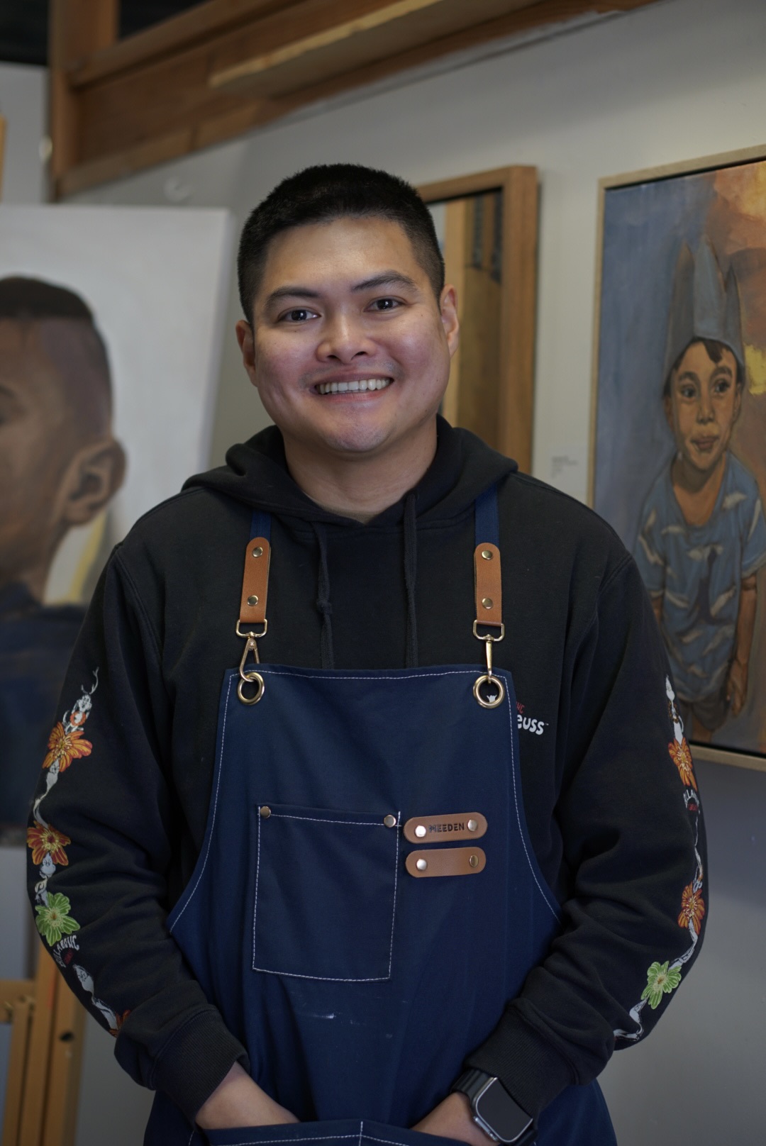Young man smiling, wearing a black hoodie and blue apron, standing indoors with artwork on the wall behind him.