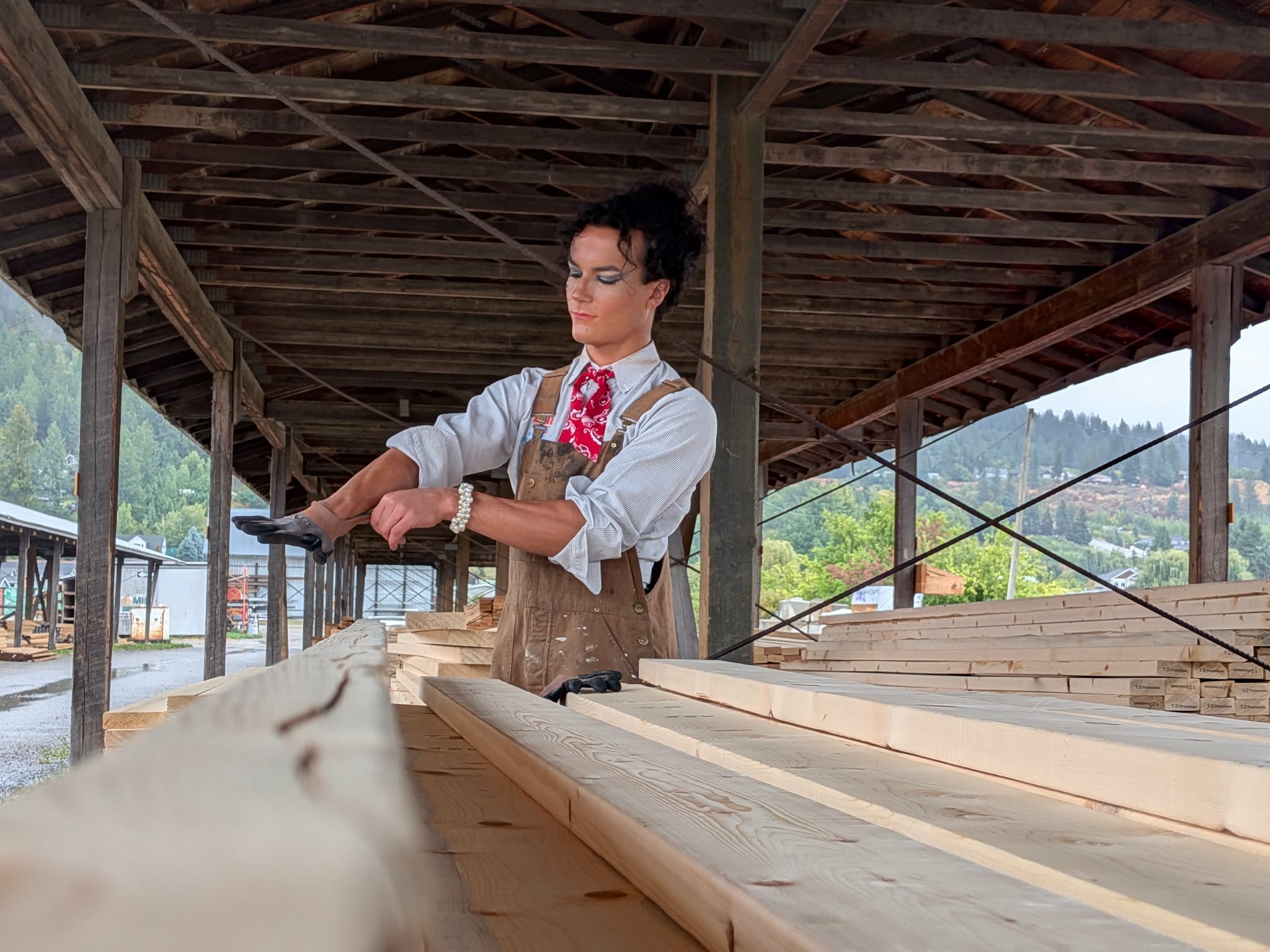 Woman working with wooden planks in a construction site under a wooden roof, outdoors with trees and mountains in background.