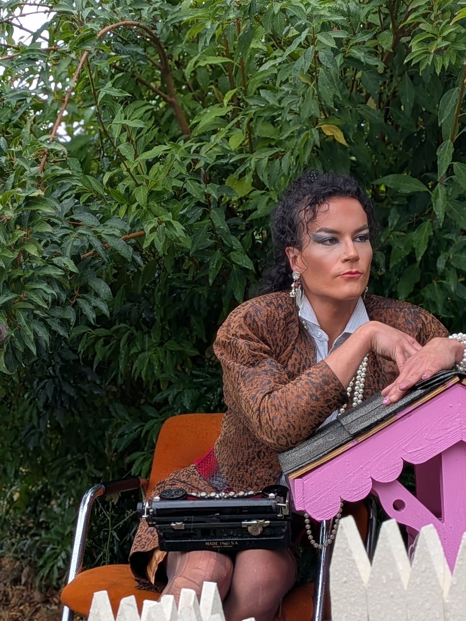 Woman with short curly hair and earrings, wearing a brown jacket, sitting outdoors with green foliage in background, resting her chin on her hand.