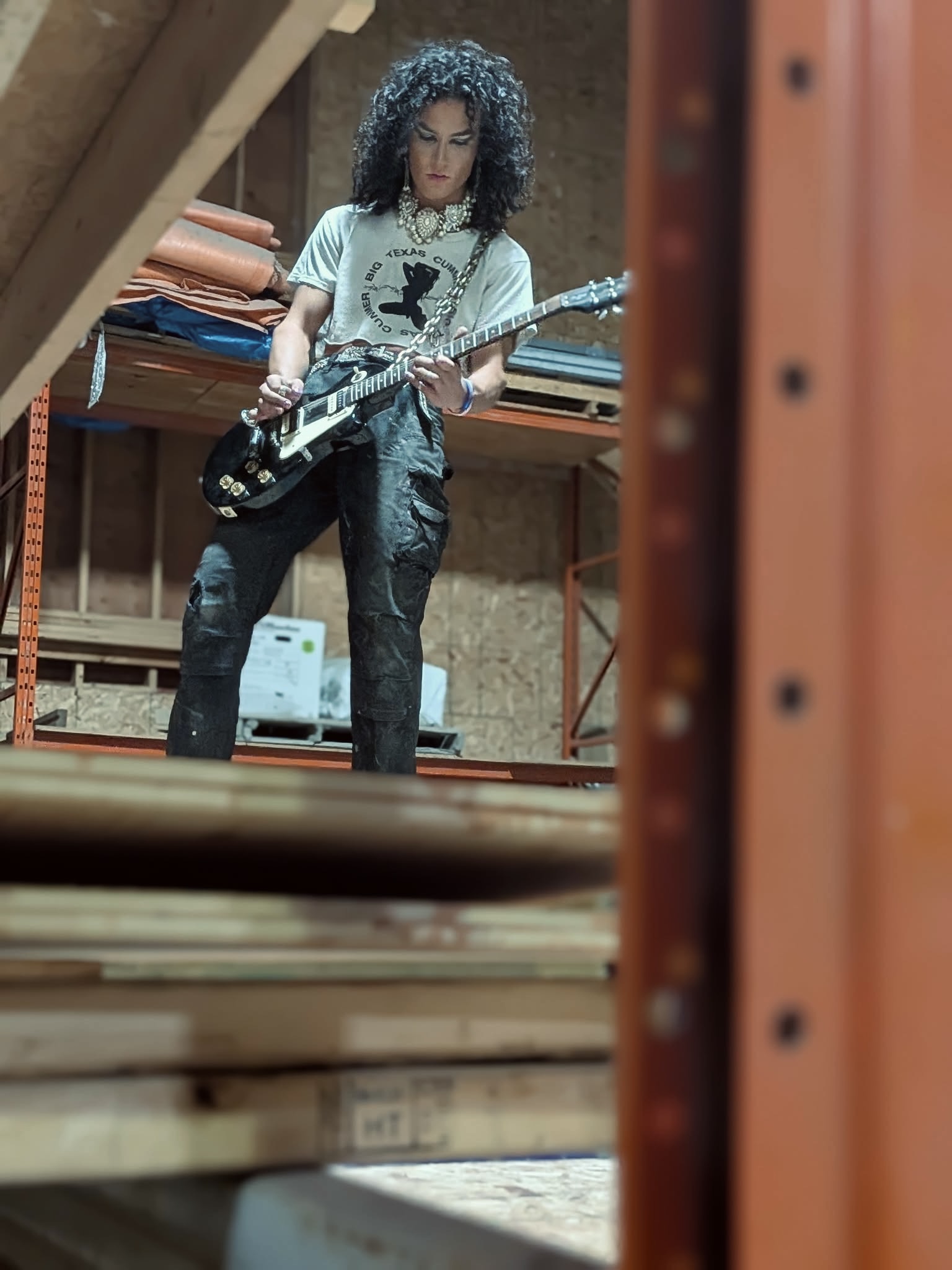 Young woman with curly hair playing an electric guitar in a storage room with shelves and wooden planks.
