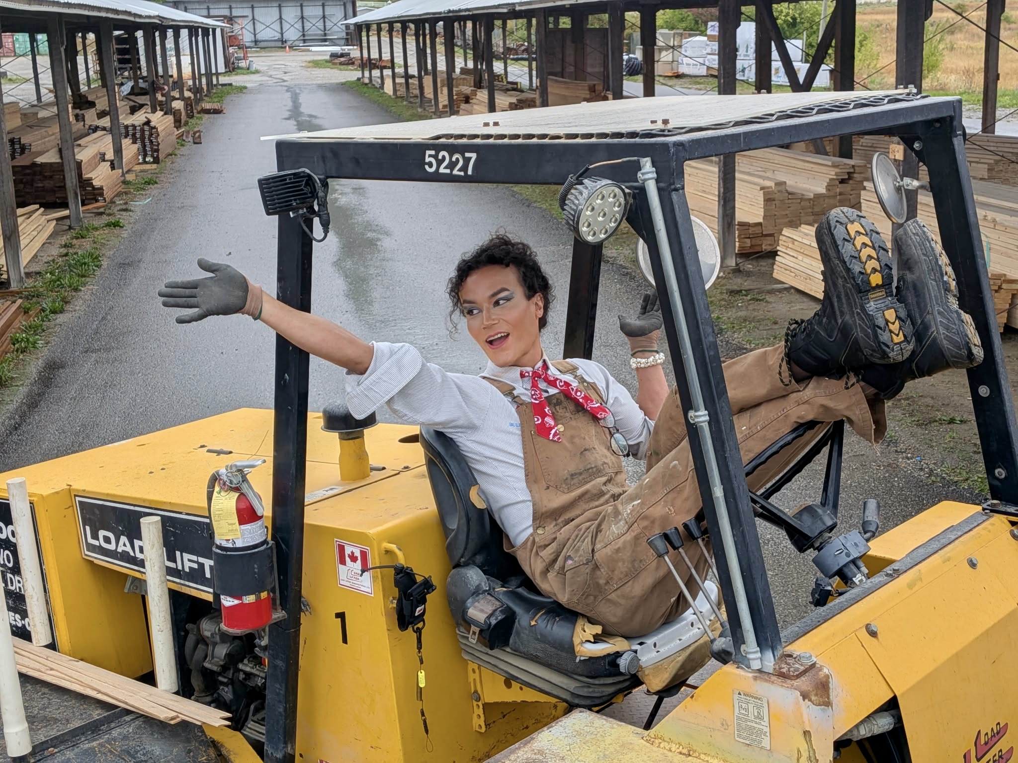 Woman sitting in a yellow forklift, smiling, with one arm extended, outdoors on a rainy day, surrounded by wooden pallets.
