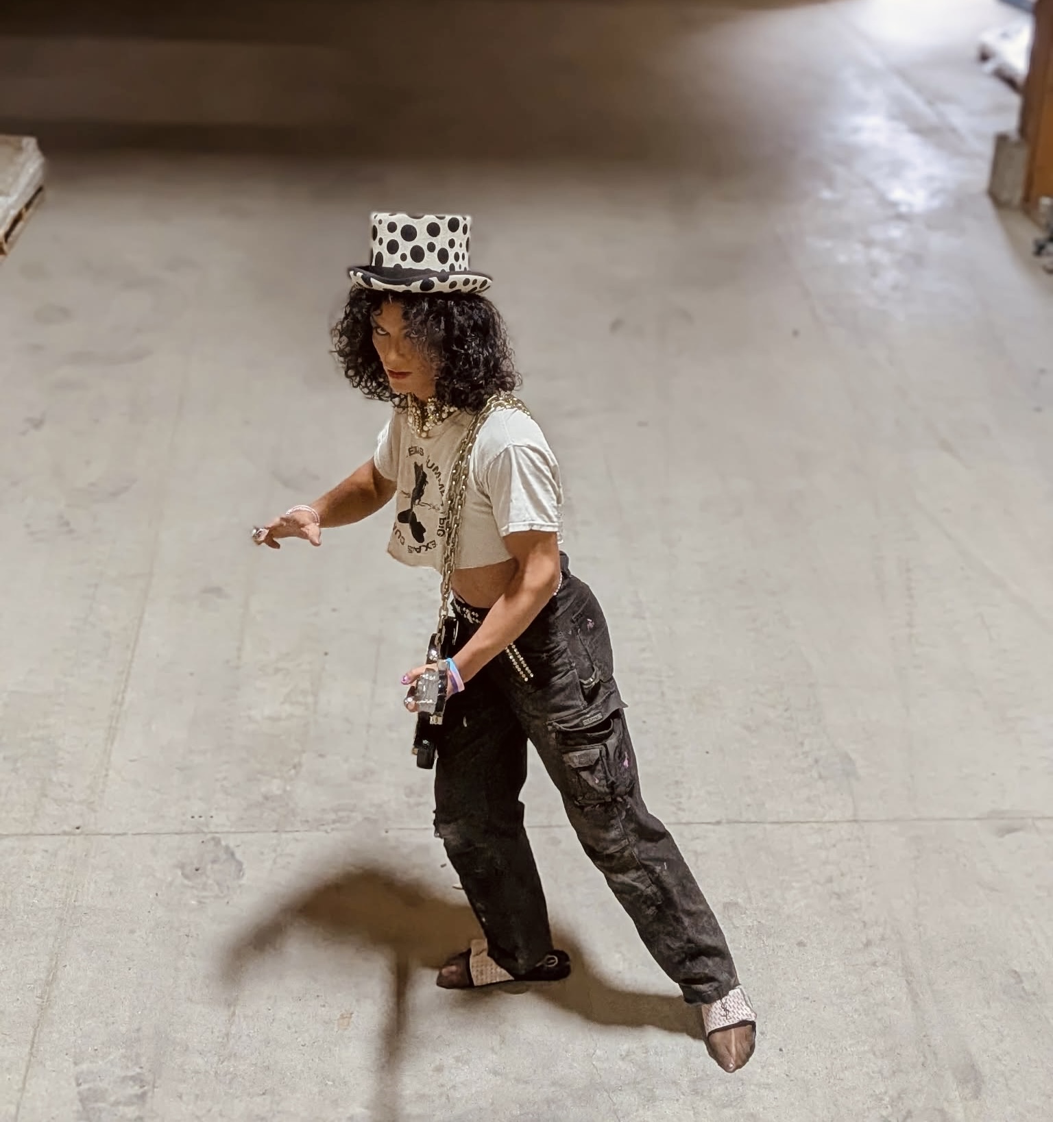 Child wearing a polka dot hat, white shirt, and black pants, standing on a concrete floor, looking down, holding an object.