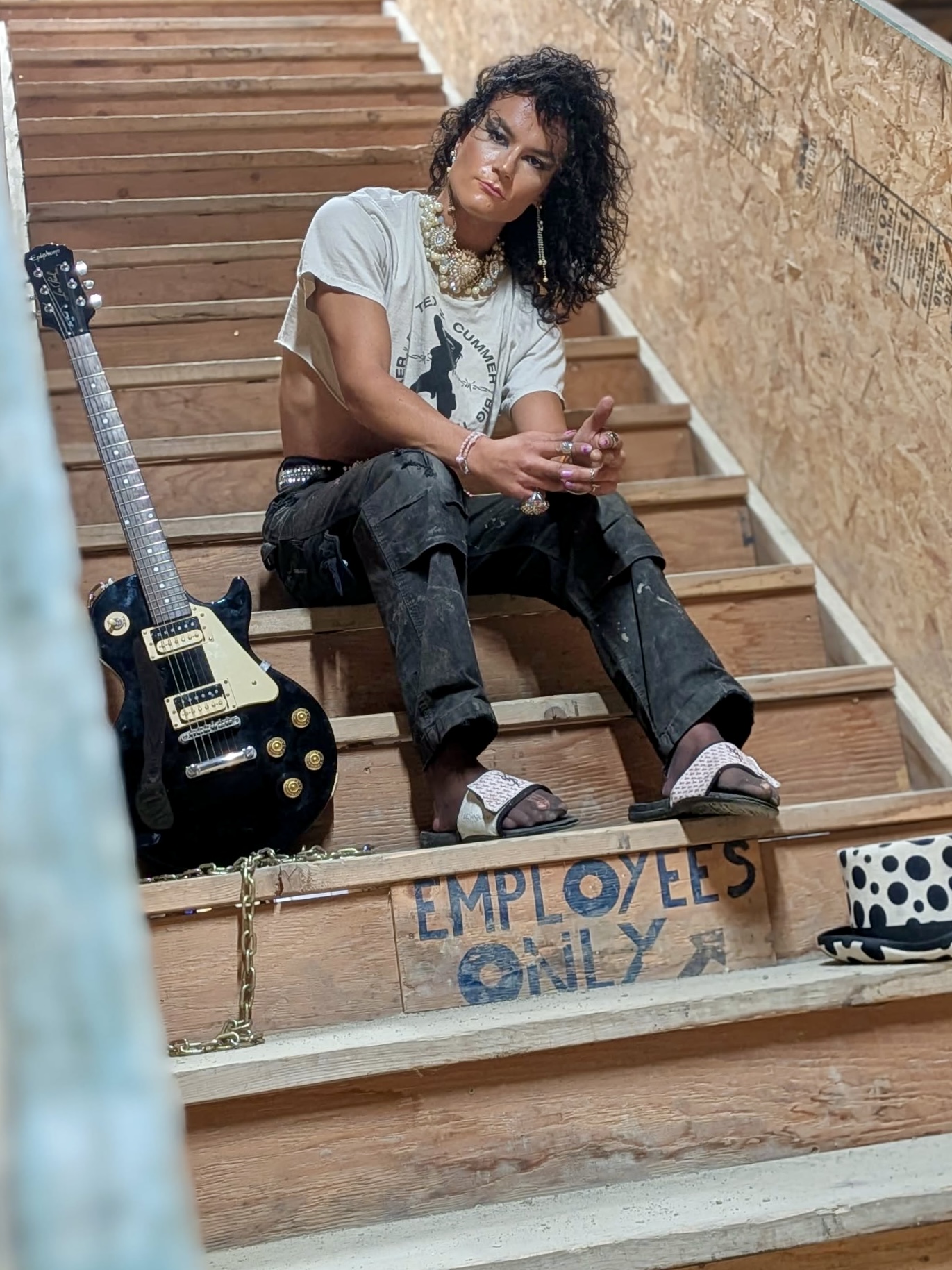 Woman sitting on stairs with a guitar, wearing a light shirt, dark pants, and sandals, holding a phone, with a sign reading 'Employees Only' nearby.