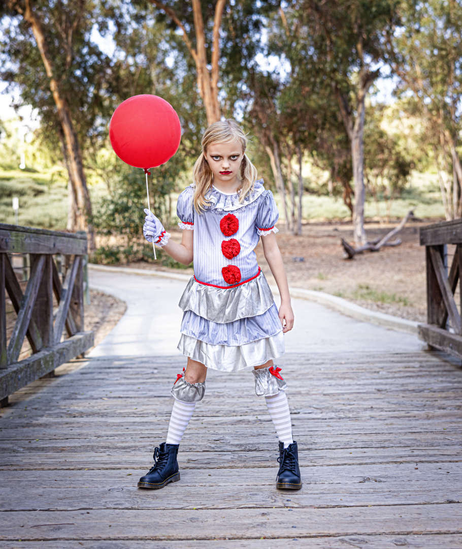 Girl dressed as a clown holding a red balloon on a wooden bridge, trees in background.