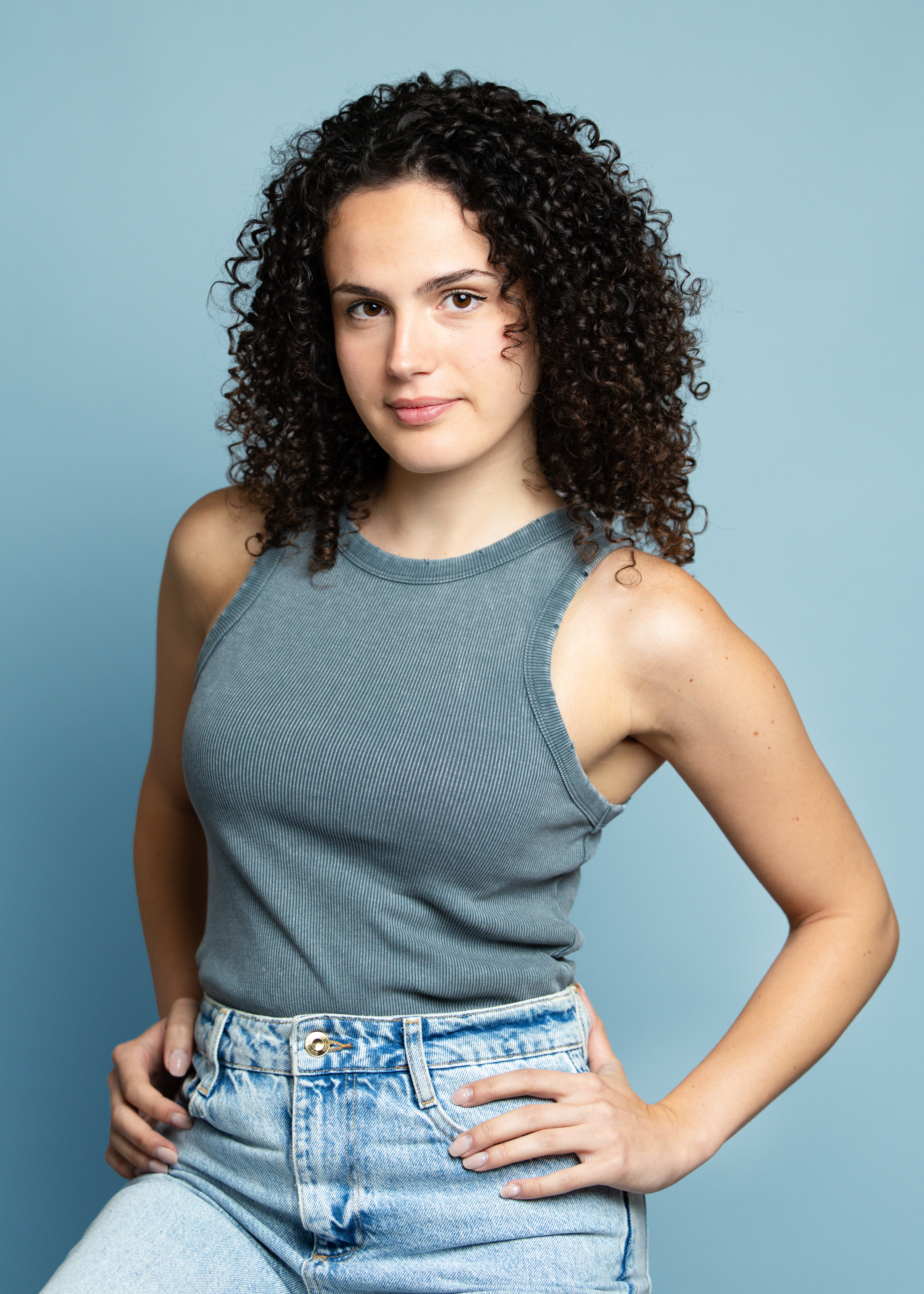 Young woman with curly hair wearing a sleeveless gray top and jeans, standing with hands on hips against a light blue background.