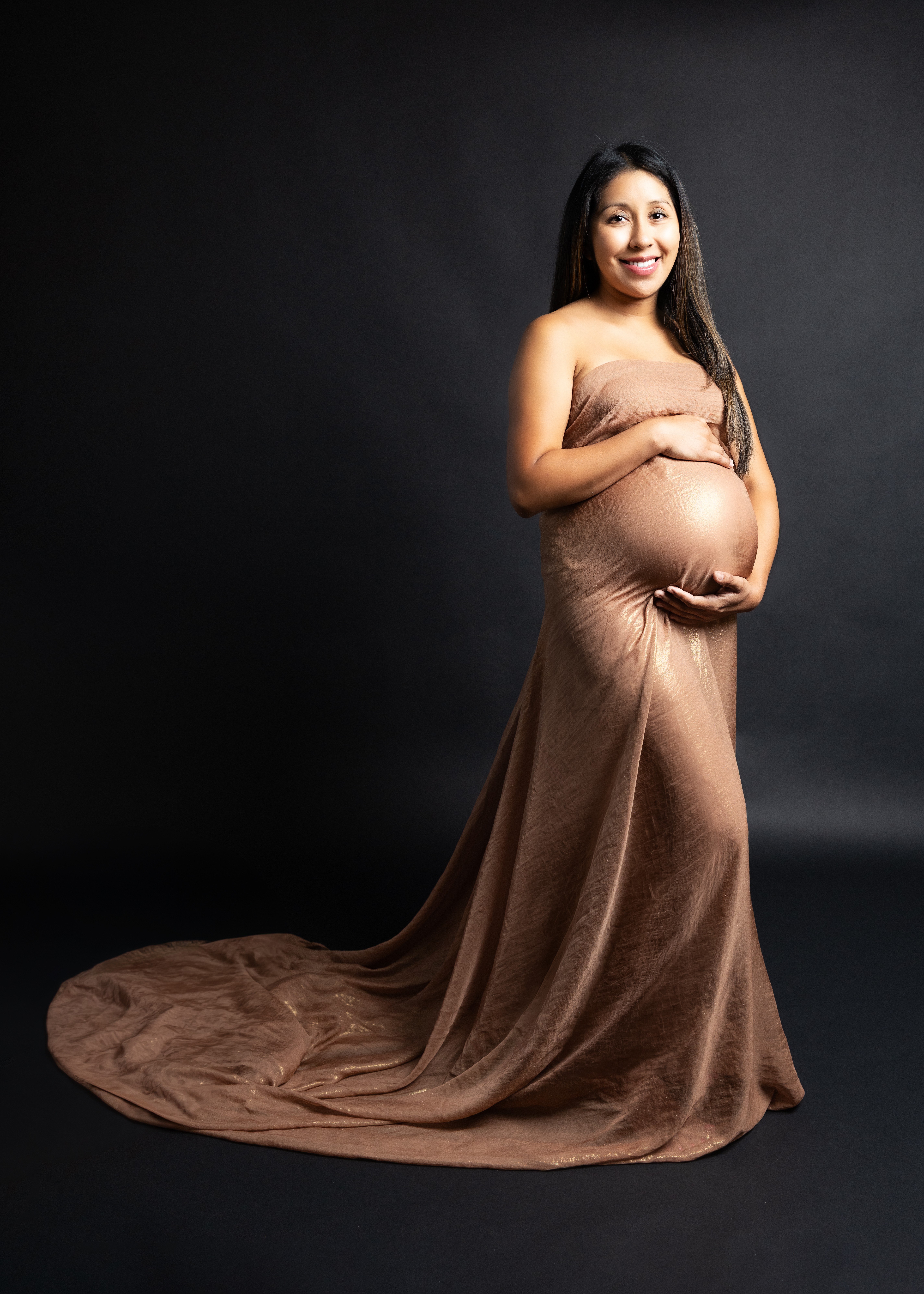 Pregnant woman in a brown gown holding her belly, standing against a dark background.