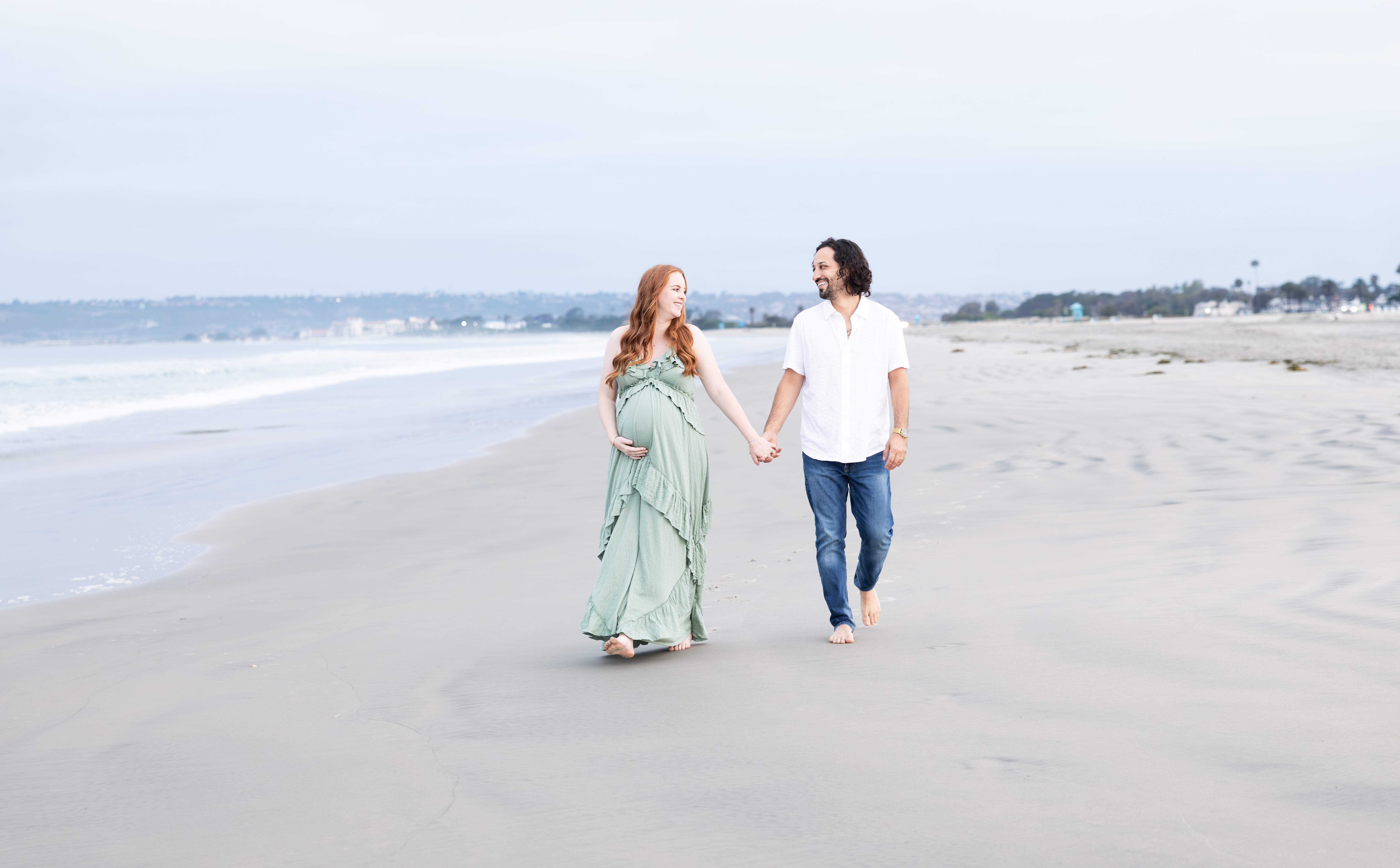 A couple holding hands walking on a sandy beach with ocean and sky in the background.