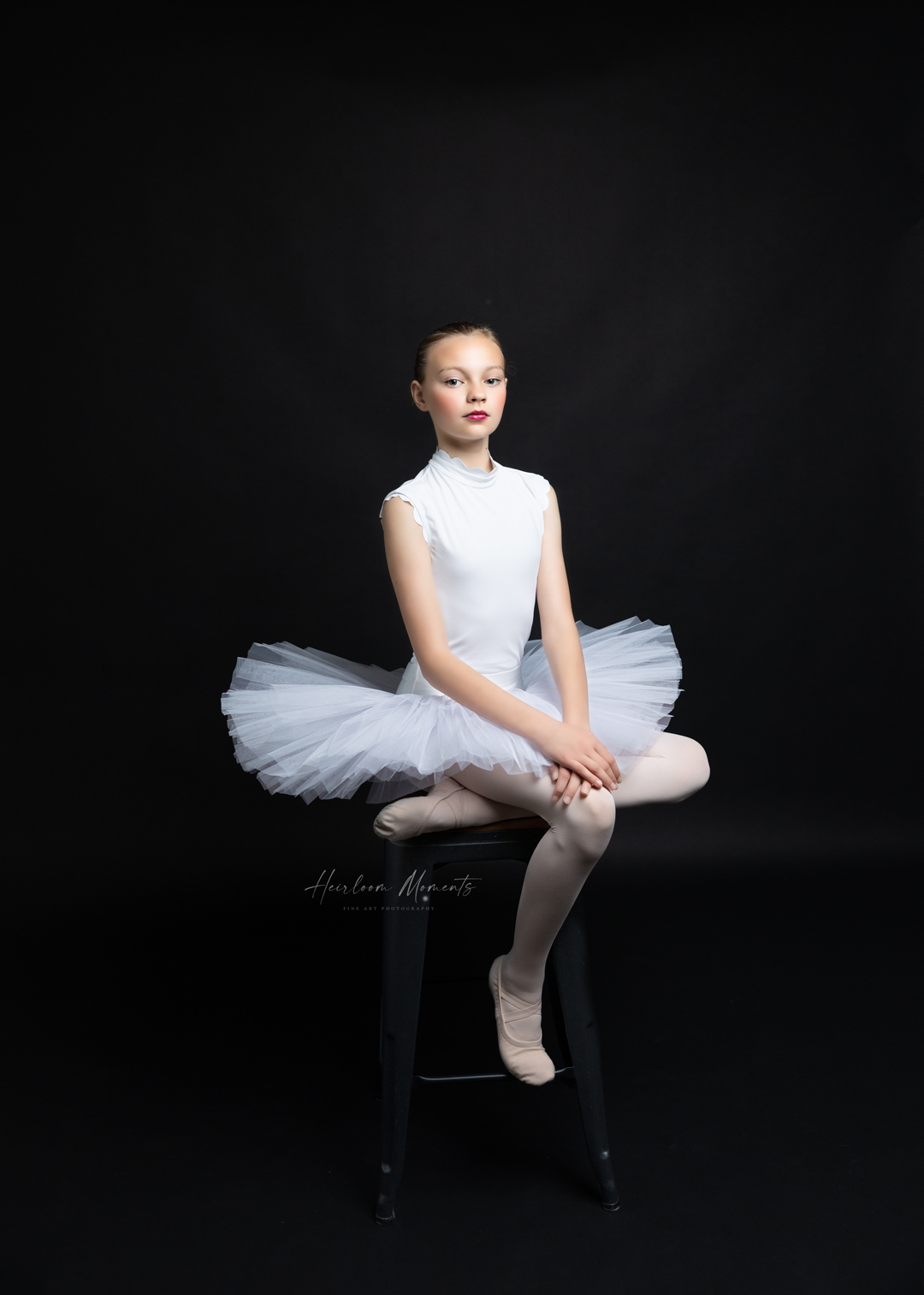 Ballet dancer in white tutu and tights sitting on a black chair against a dark background.