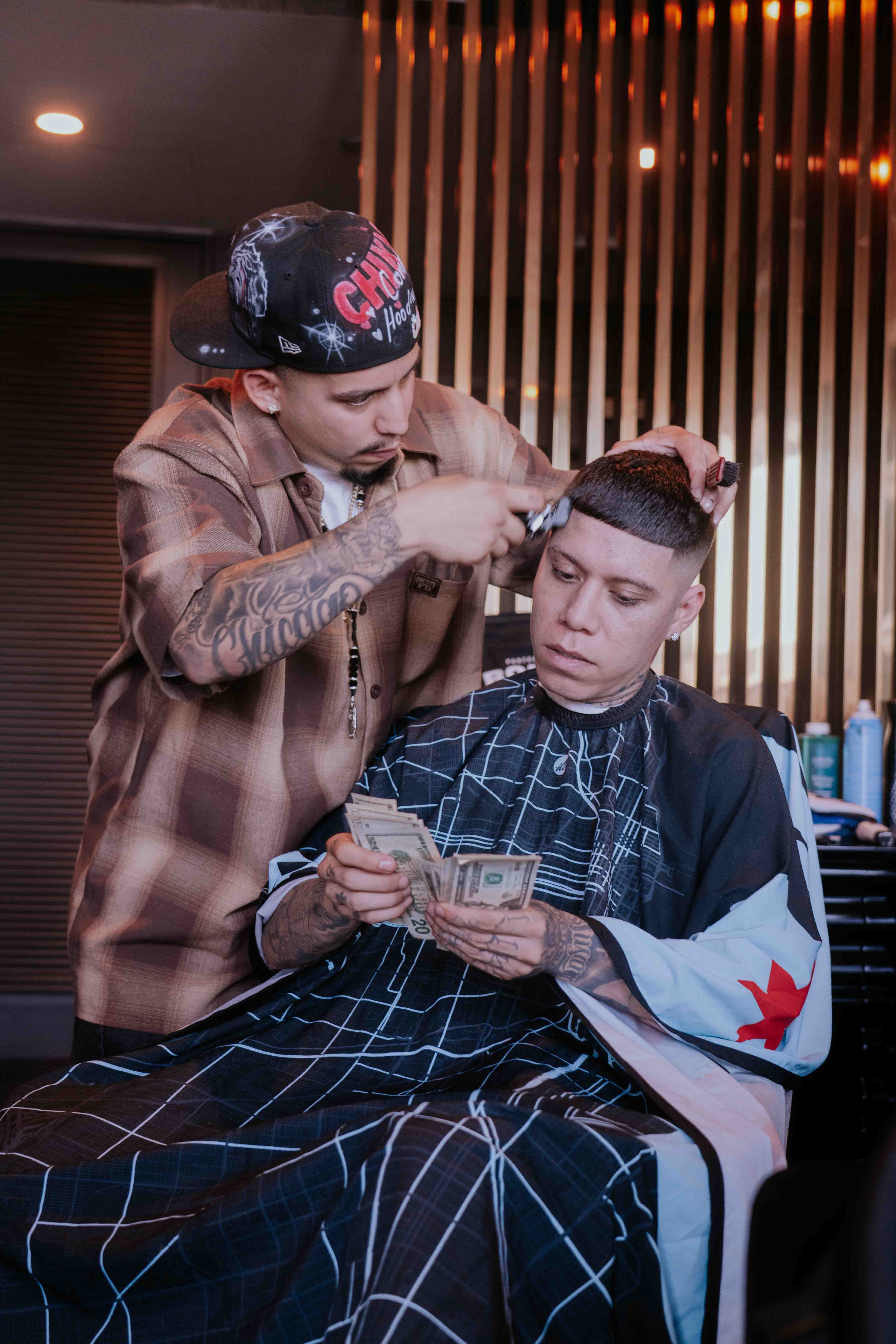 Barber cutting young man's hair in a salon with wooden wall decor, young man reading a phone, barber wearing a cap, tattooed arm visible.