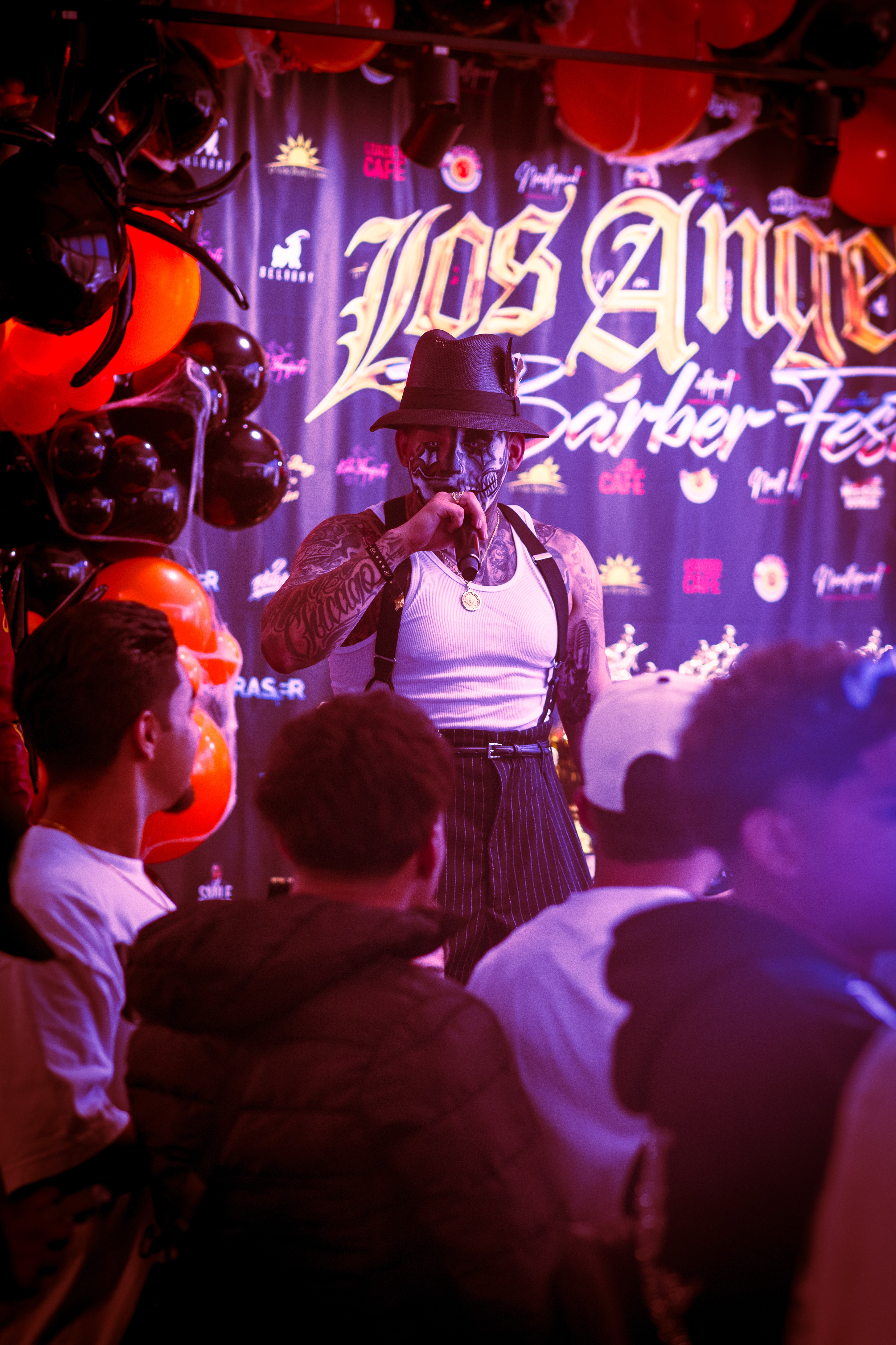 Performer with a hat and microphone on stage, audience watching, colorful balloons, and a backdrop with 'Los Angeles' text.