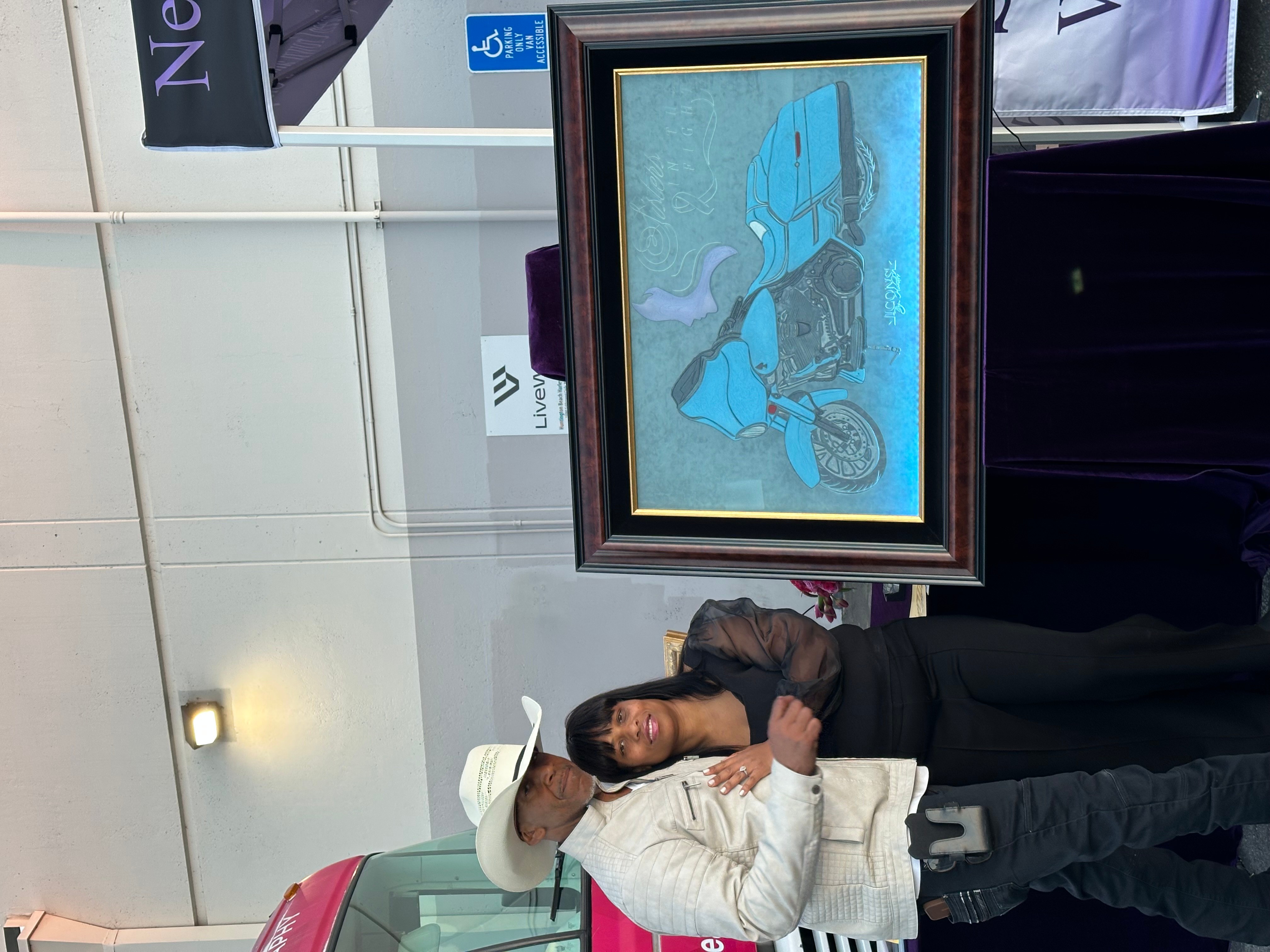 Woman in white coat and hat standing next to framed artwork of a blue horse, in an indoor setting.