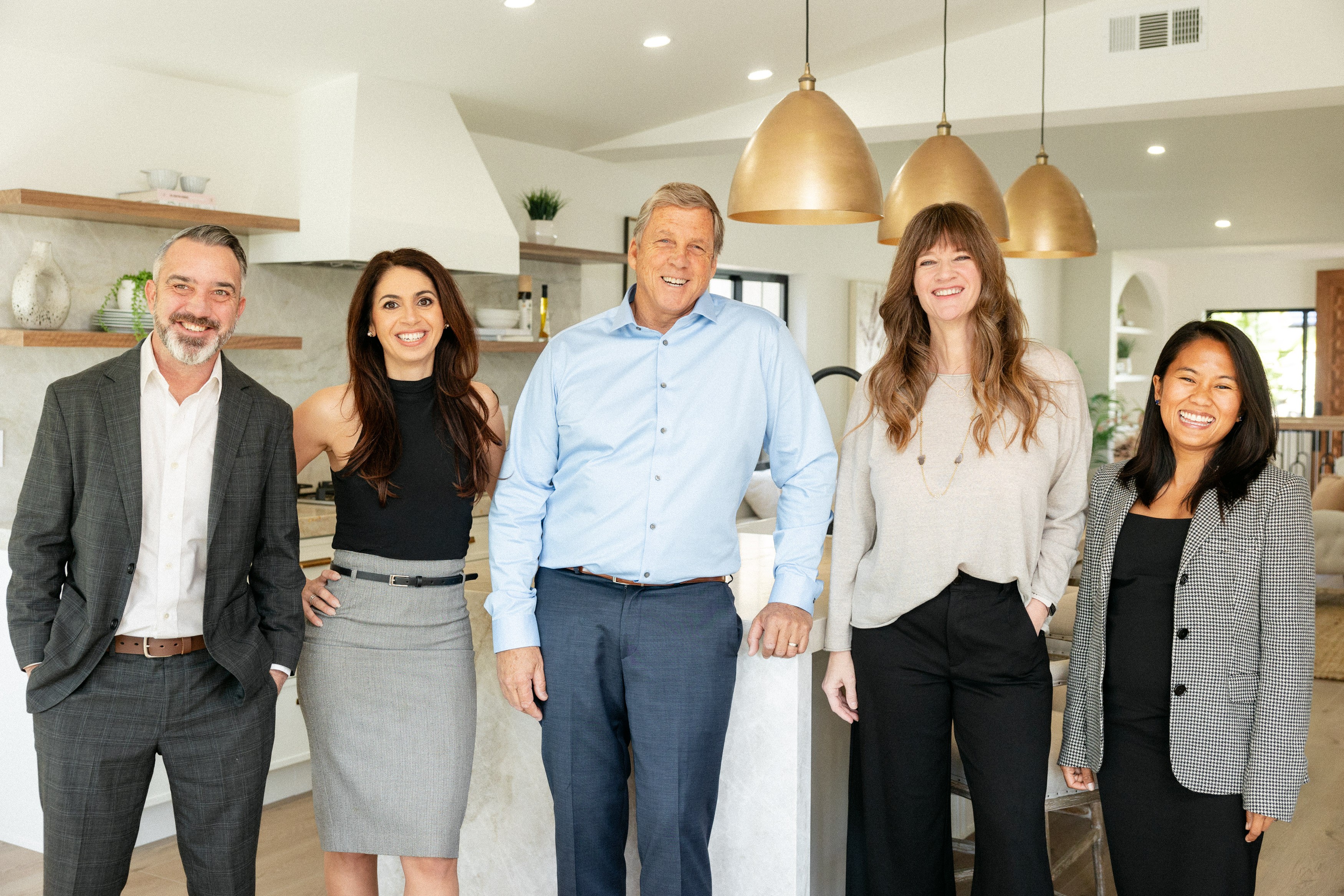 Group of five diverse professionals standing in a modern kitchen, smiling, with pendant lights overhead.