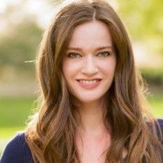 Young woman with long brown hair smiling outdoors in a park, wearing a dark top.