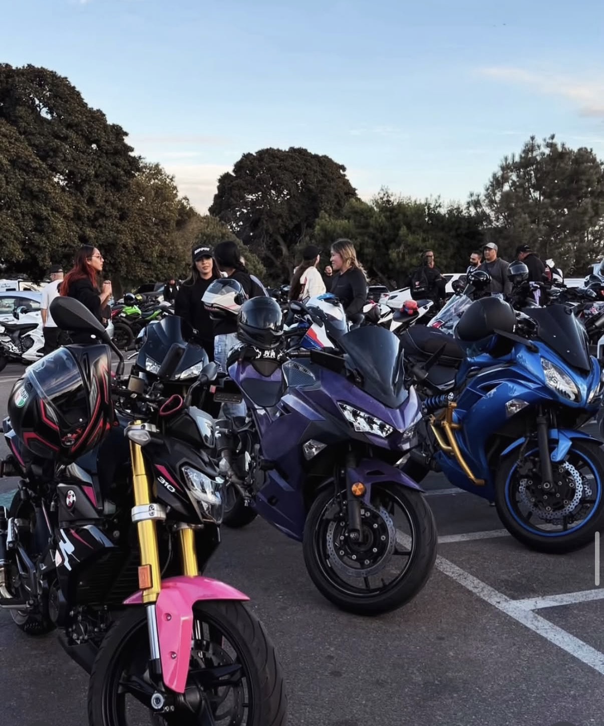 Motorcycles parked in a lot with people standing nearby, trees in the background, clear sky.