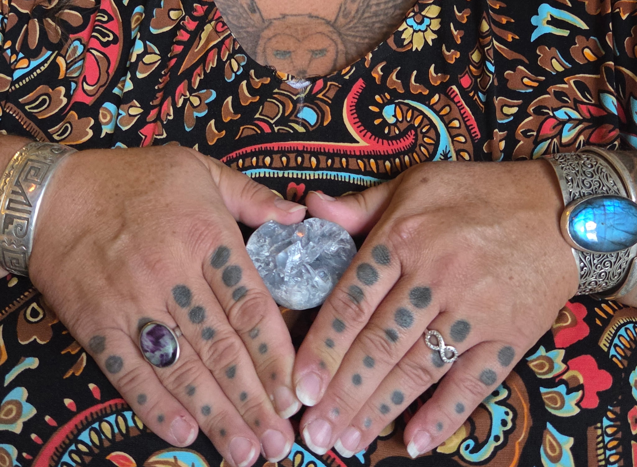 Person's hands holding a clear crystal, with tattoos and rings, on a colorful patterned fabric background.