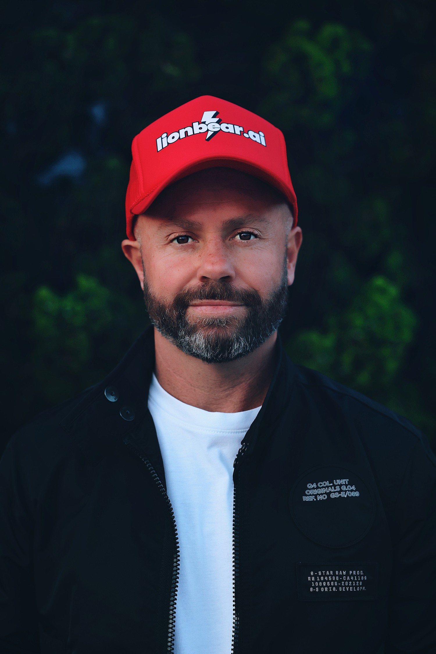 Man with beard wearing a red cap and black jacket outdoors, dark green background