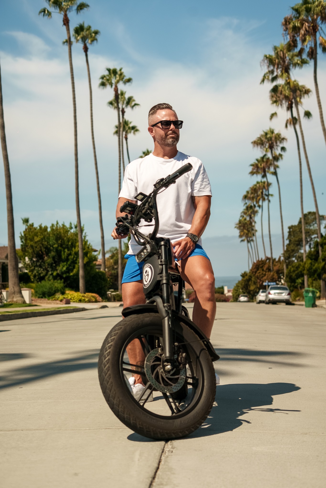Man sitting on a black electric bike on a sunny street with tall palm trees in the background.