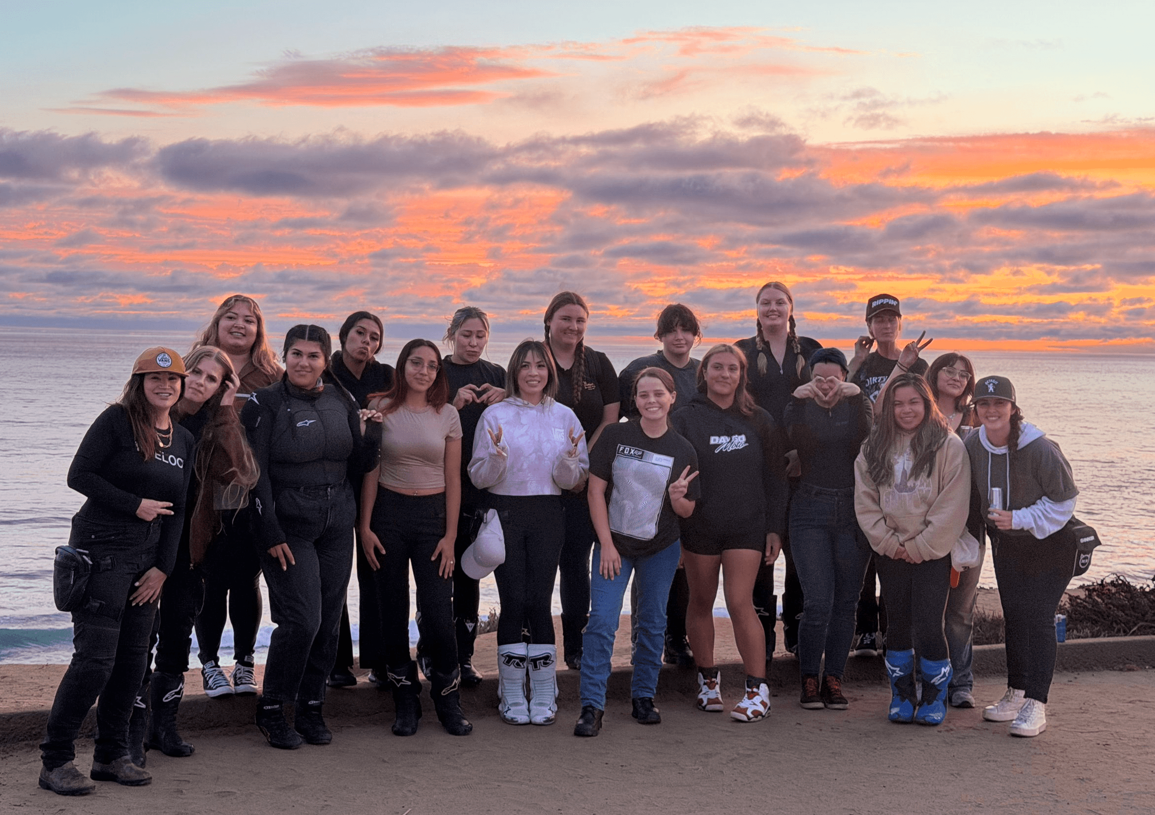 Group of people standing on a beach at sunset, with colorful sky and ocean in background.