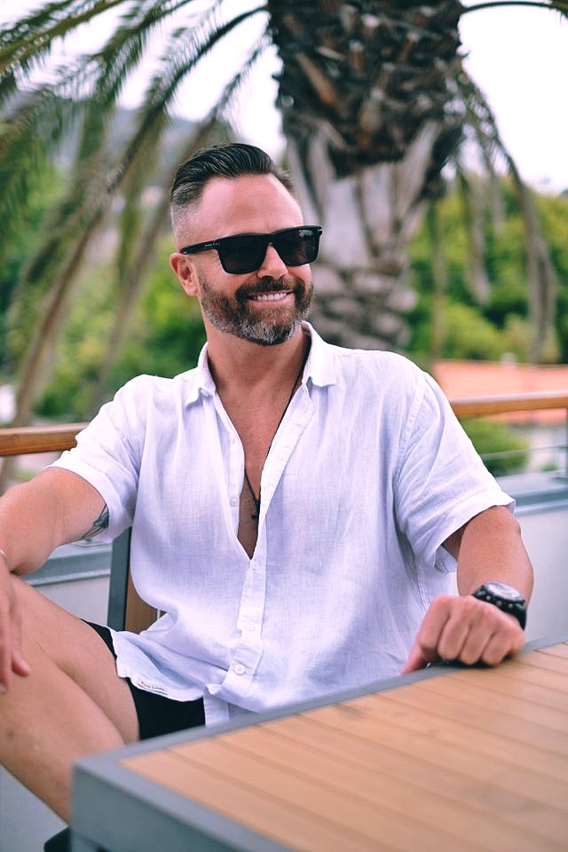Man with sunglasses and a beard smiling outdoors, wearing a white shirt, sitting at a table with greenery in background.