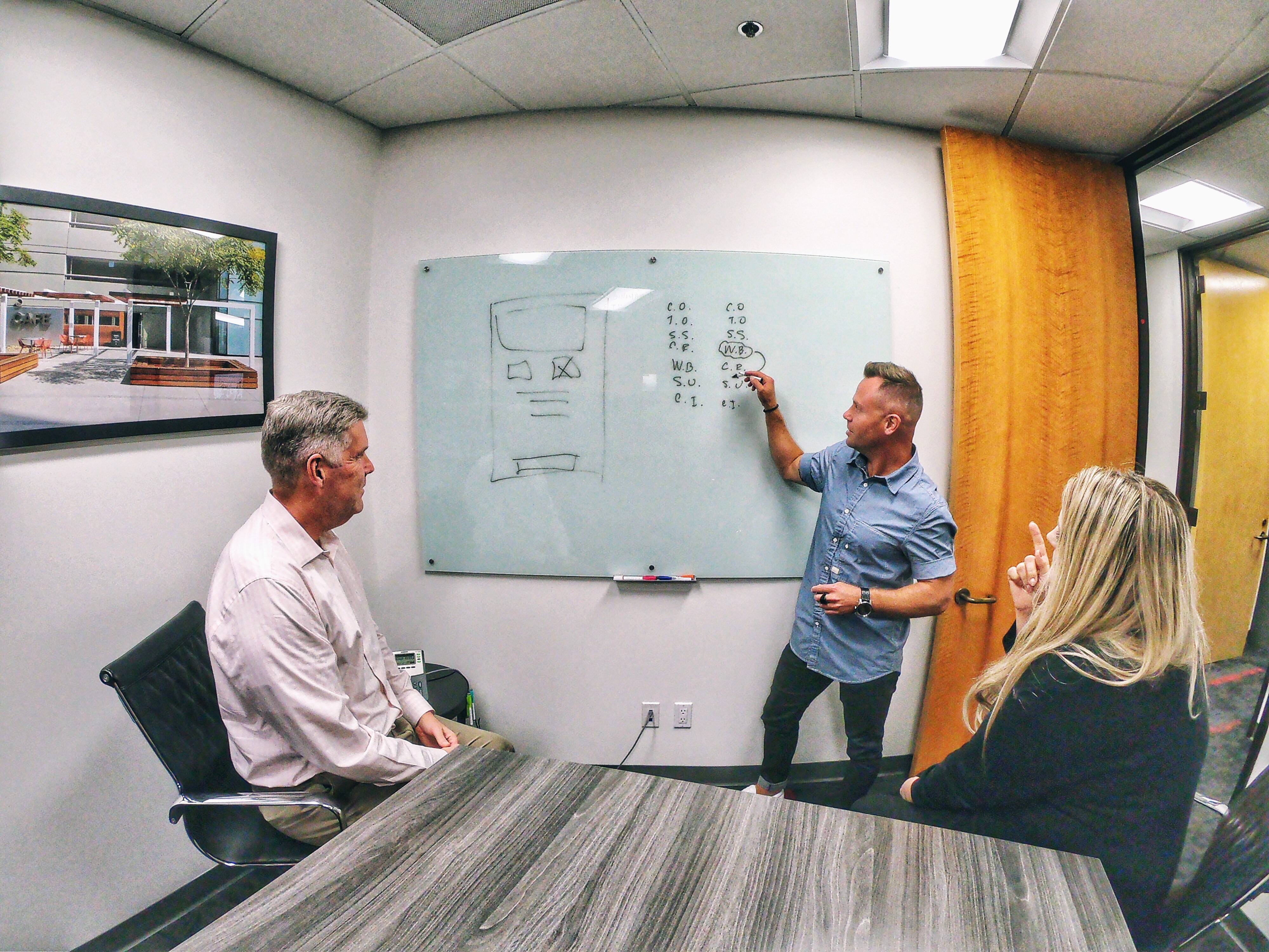Three people in a meeting room, one person standing and writing on a whiteboard, two seated listening, a window with a view outside.