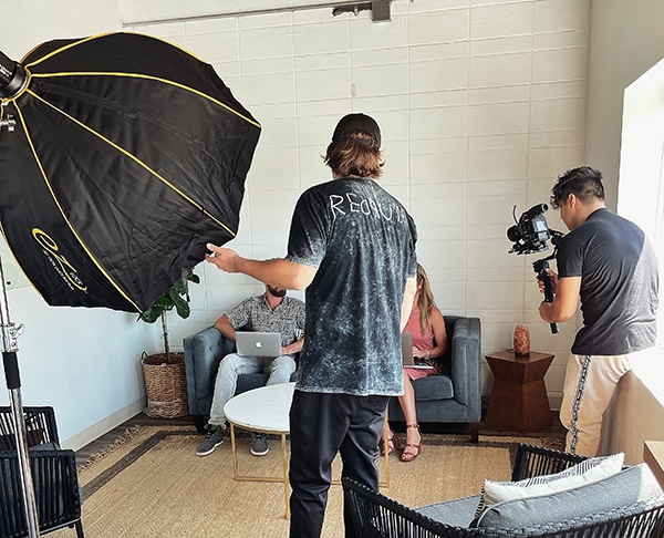 Person standing in a room with a camera operator filming, others seated on a sofa, large black umbrella for lighting, and a white wall.