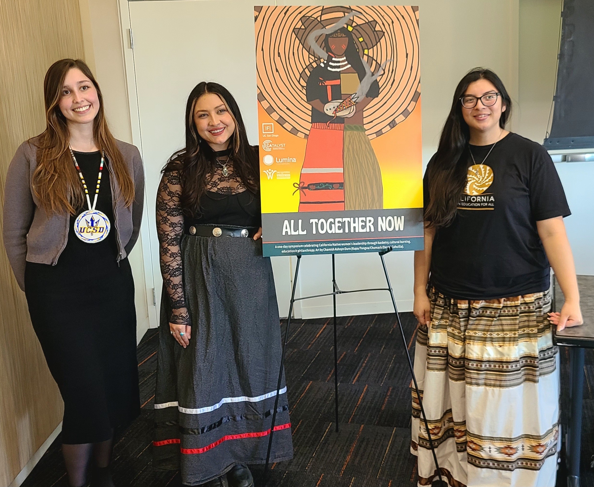 Three women standing indoors next to a colorful poster on an easel, smiling, with a whiteboard behind them.